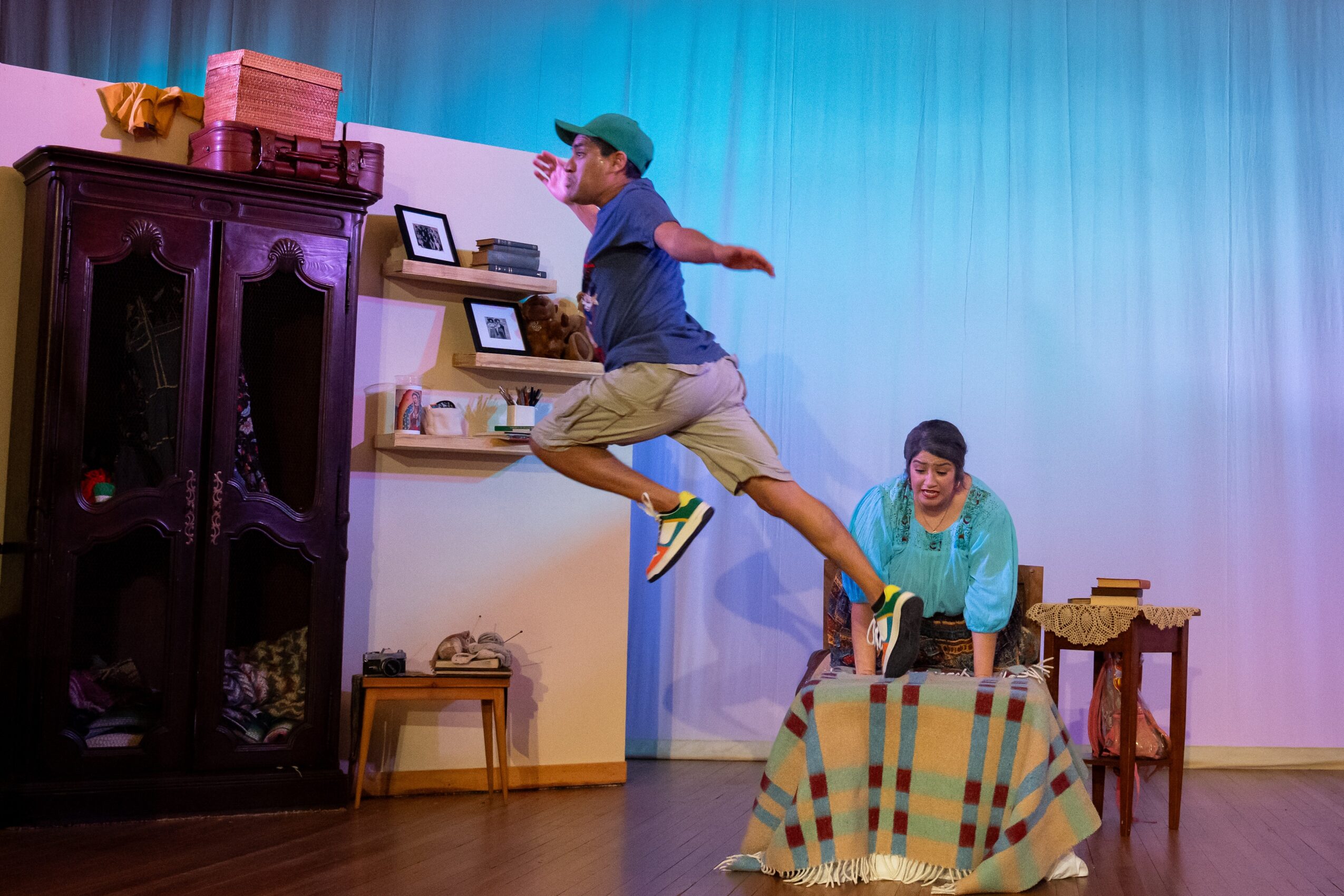 A person leaps beside a table as another person sits on a stage set with a wardrobe and shelves.