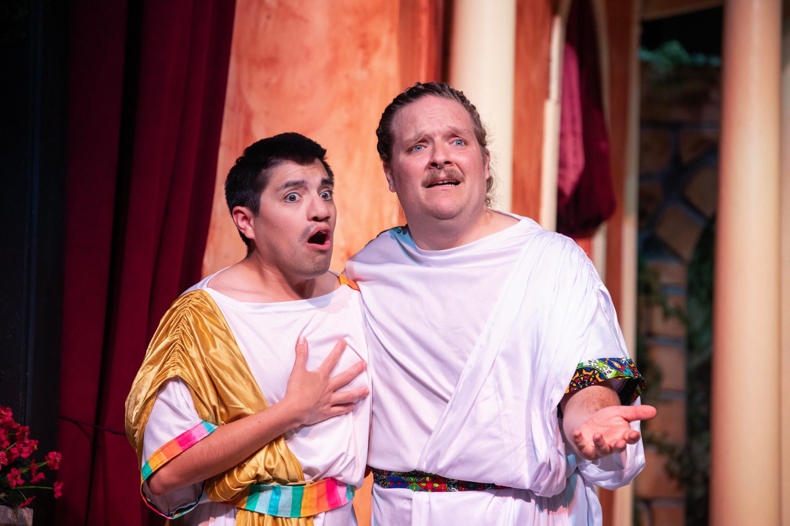 Two people in theatrical costumes stand onstage, one with a hand on their chest and the other gesturing outward.