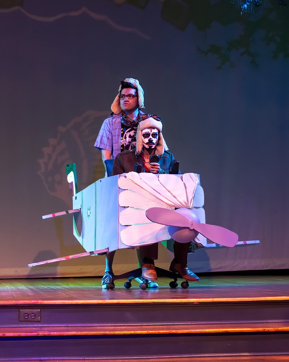 Two people in costumes stand inside a cardboard airplane prop on a stage.