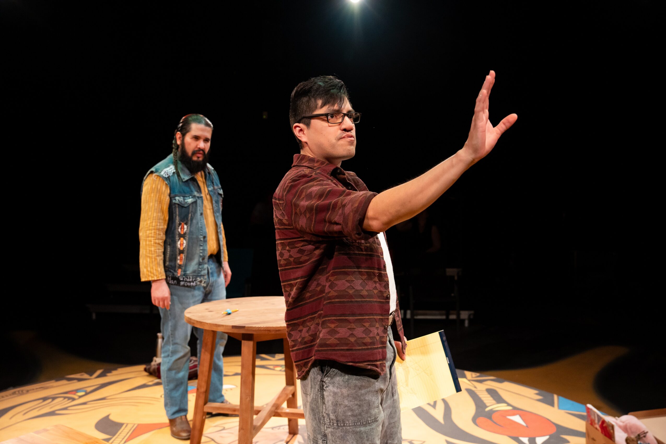 Two people stand on a stage; one raises a hand beside a wooden stool.