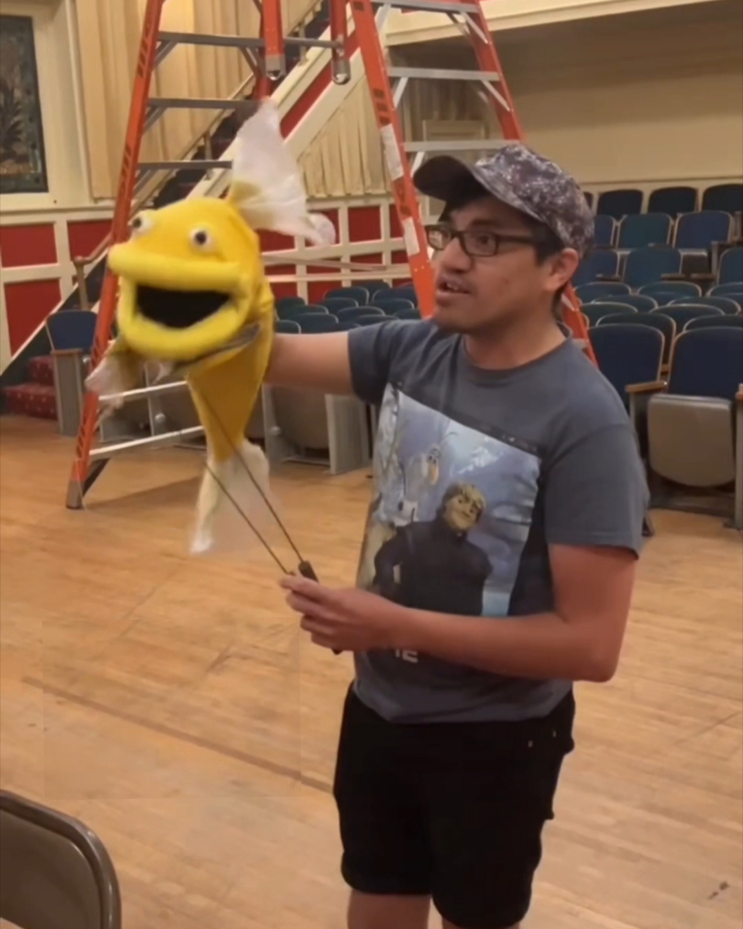 A person holds a yellow puppet in a theater with rows of seats and a ladder behind them.