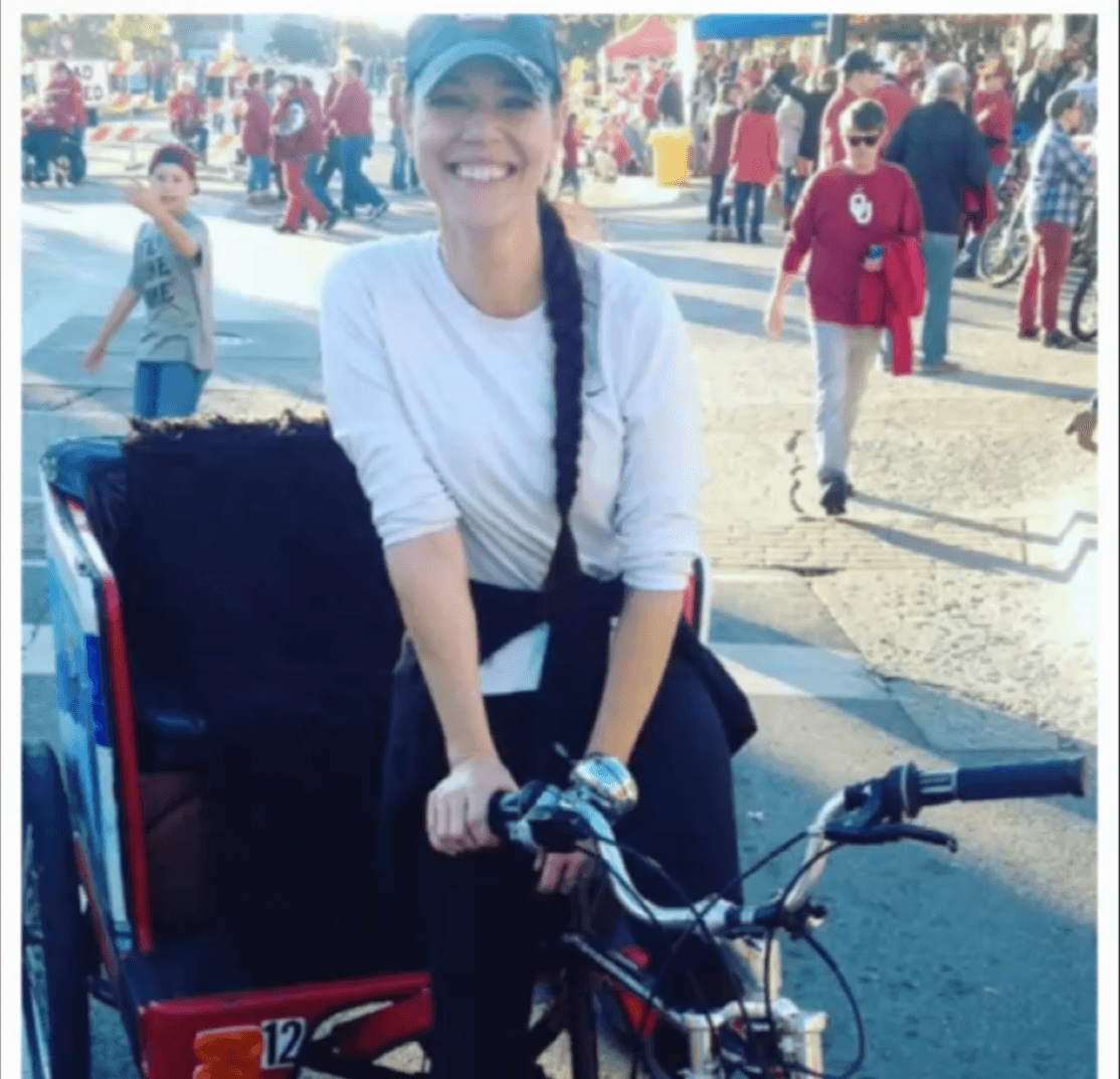 A person wearing a cap sits on a bicycle in a crowded outdoor plaza.
