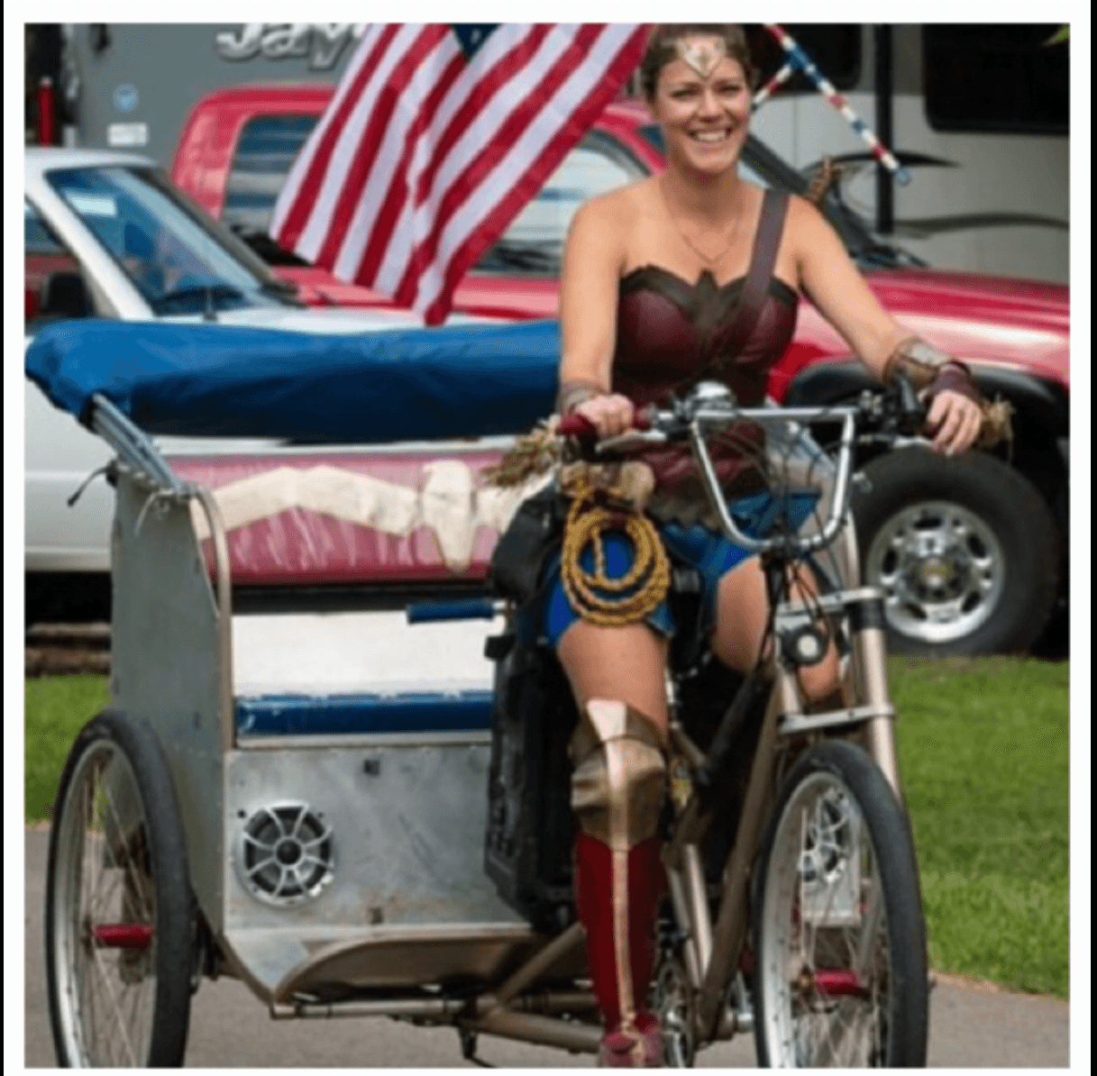 A person rides a three-wheeled bicycle with a metal cart, with parked cars and an American flag behind them.