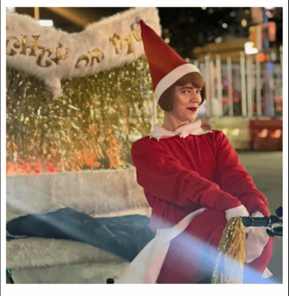 Person dressed as an elf sits on a decorated holiday float with a white banner and gold fringe.