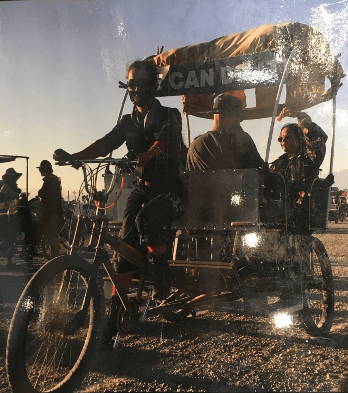 A person pedals a covered three-wheeled vehicle with passengers through a sunlit outdoor area.