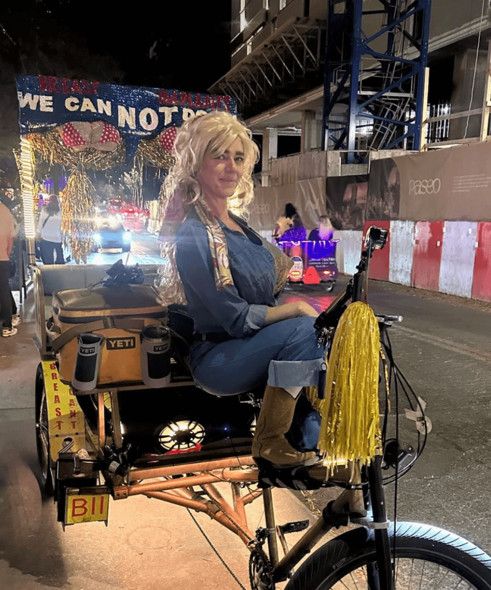 A person seated on a decorated bicycle at night, with yellow streamers and city lights in the background.