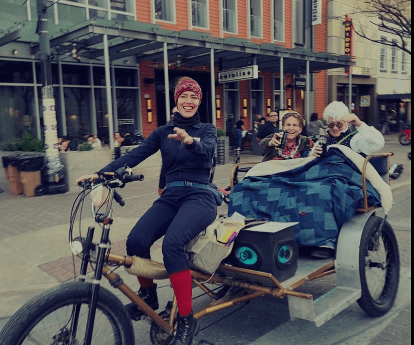 A person pedals a bicycle-powered rickshaw carrying two passengers along a city sidewalk.
