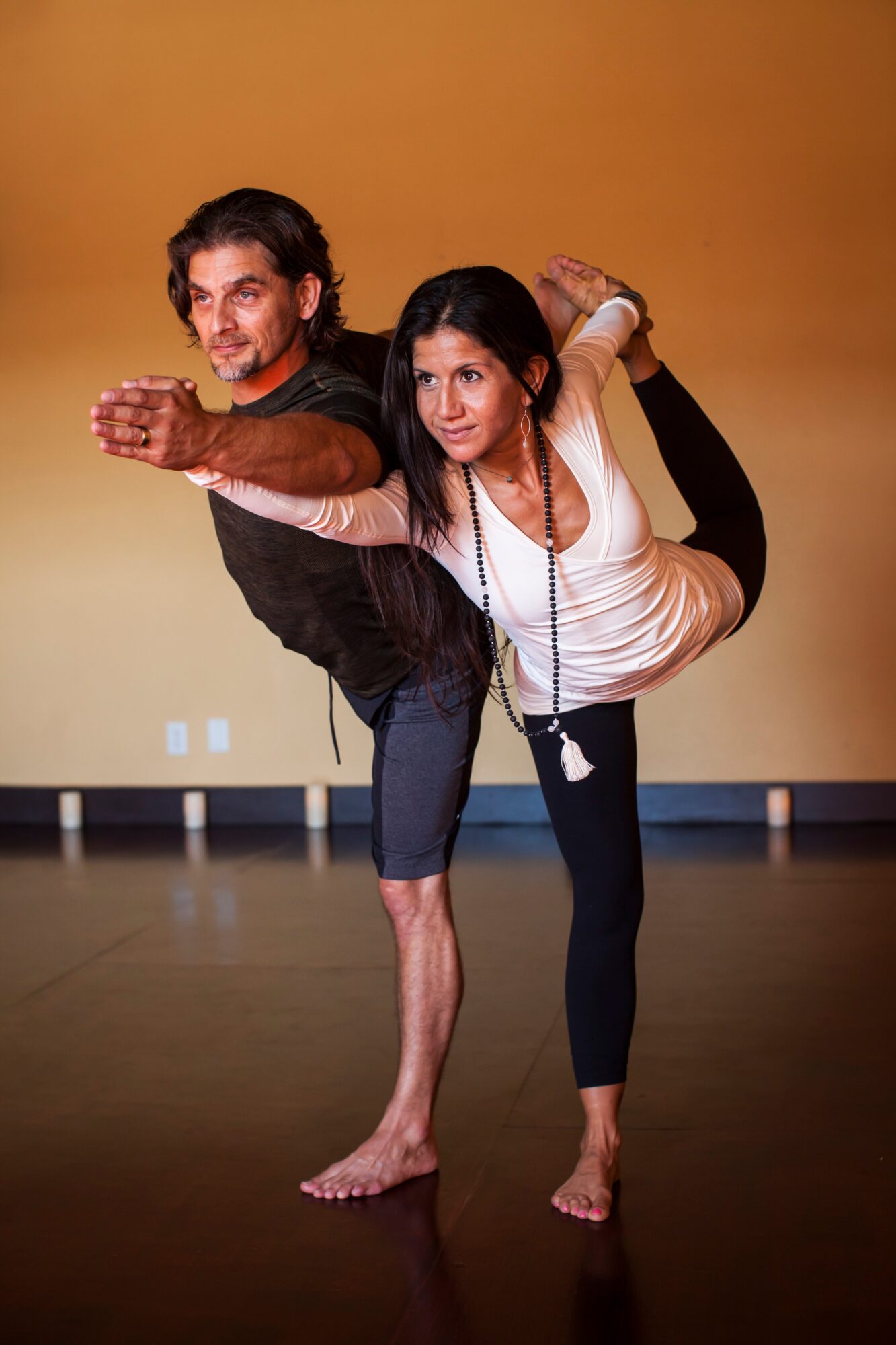 Two people balance in a partnered dance pose, leaning forward with arms extended in a studio.