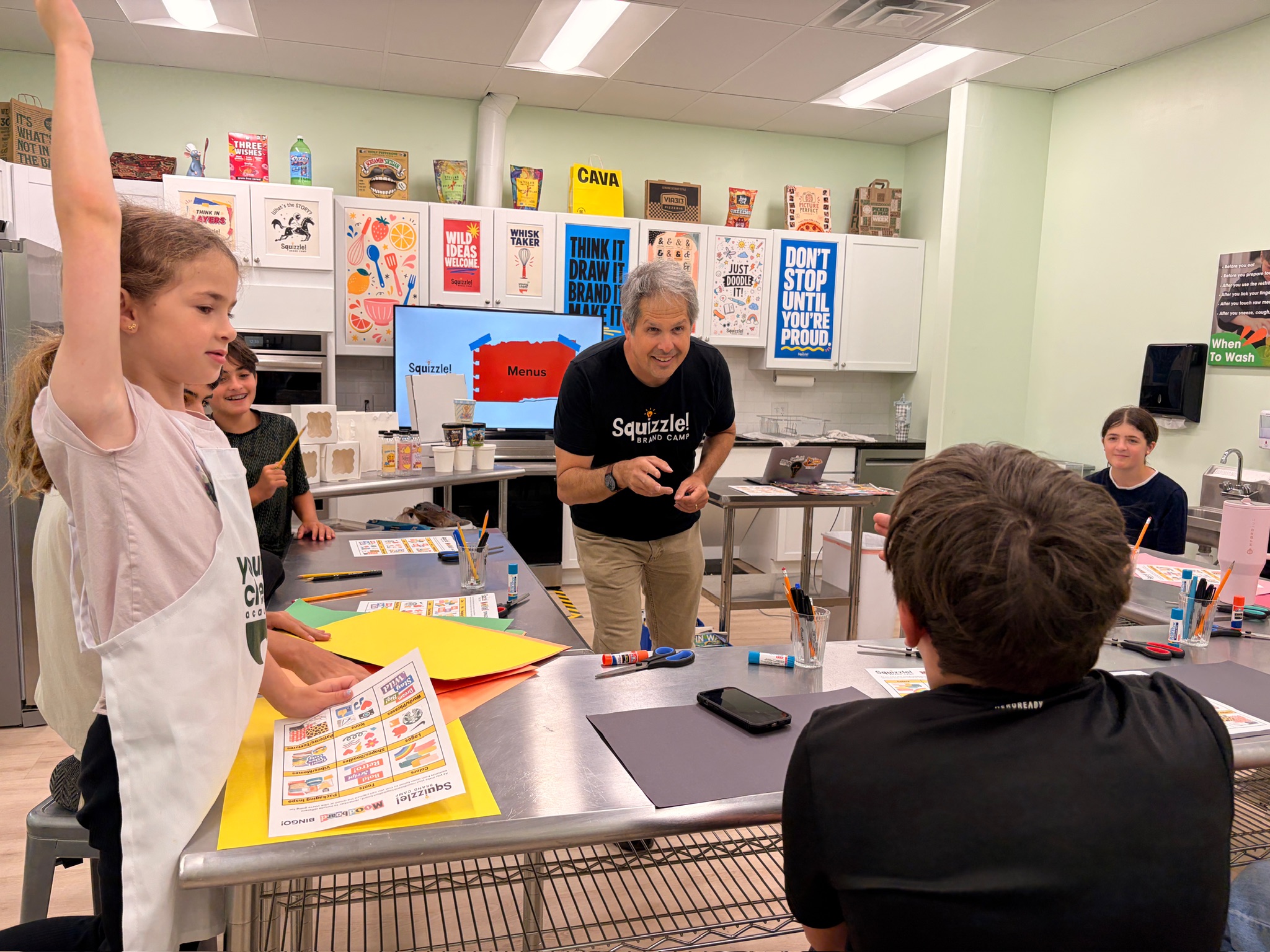 People gather around classroom tables with papers and supplies; one person raises an arm while another gestures toward the group.