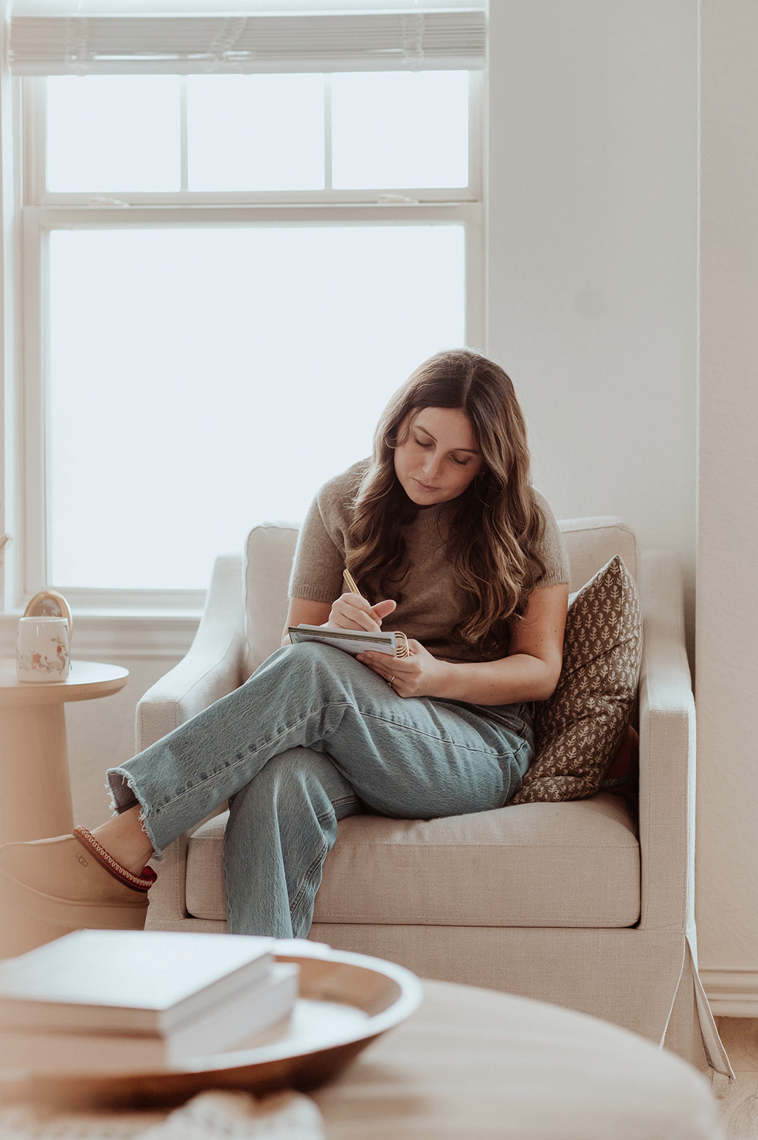 A person writes in a notebook while seated in an armchair beside a bright window.