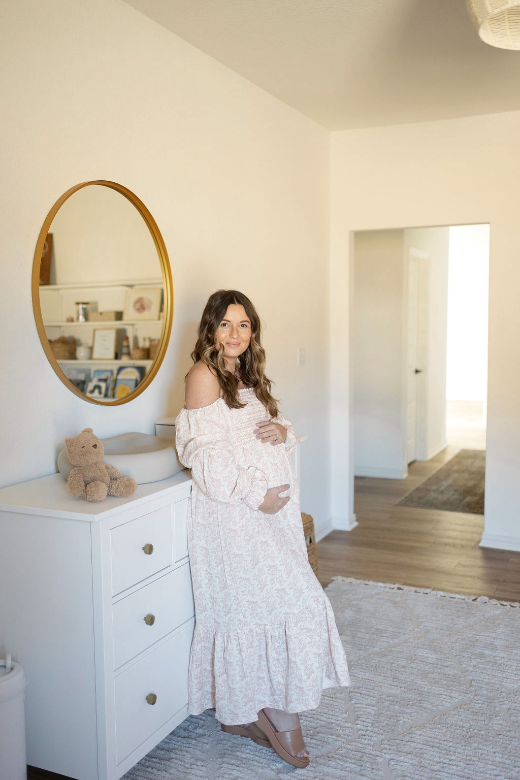 A person in a white dress stands beside a white dresser and oval mirror in a bright room.