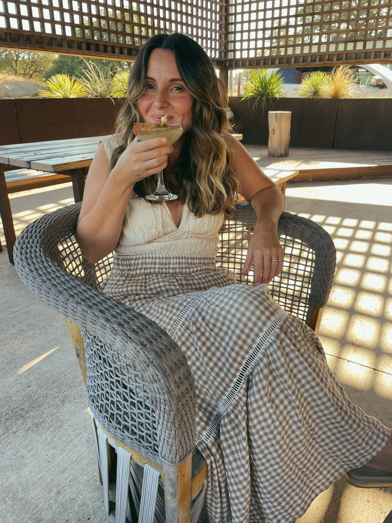 A person seated in a woven chair holds a drink and a small piece of food beneath a lattice-covered patio.