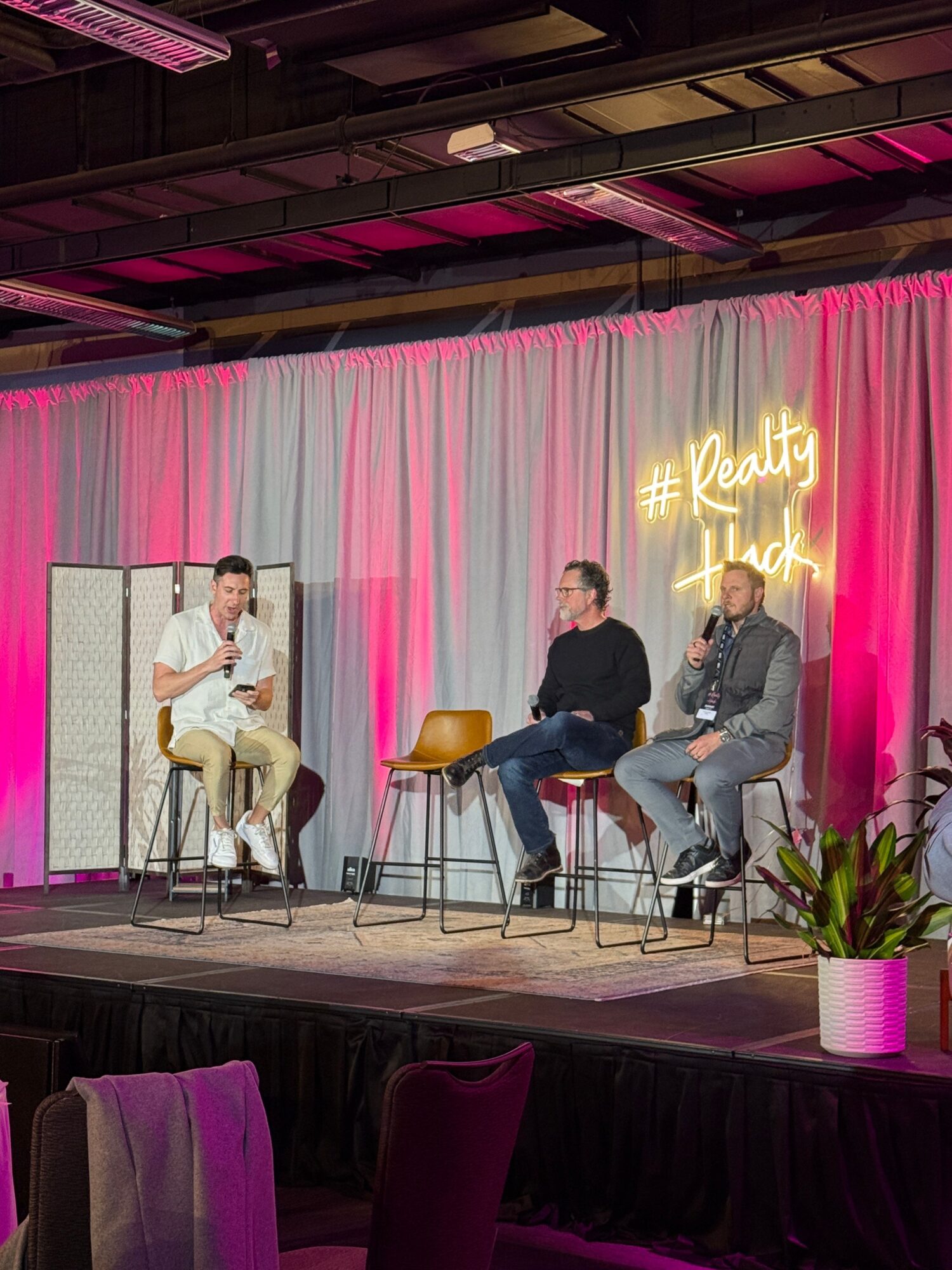 Three people seated on stools beneath pink lighting, with a glowing sign and plants behind them.