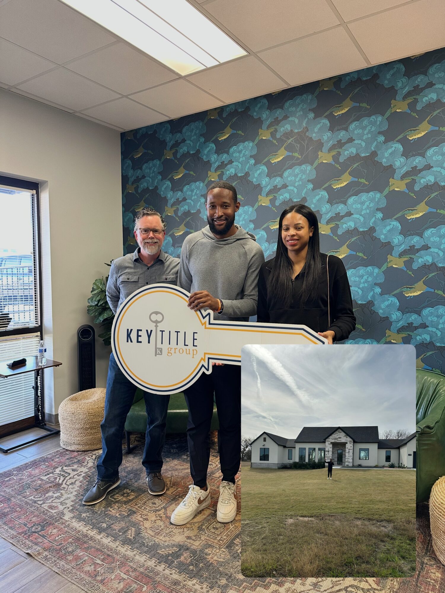 Three people pose beside a large printed photograph of a house in a room with patterned wallpaper.