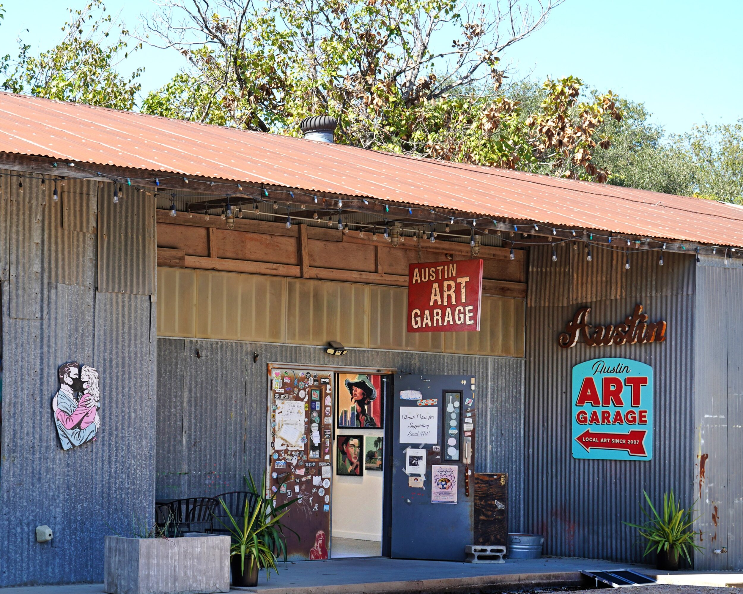 A blue corrugated-metal art garage with a red roof, wall signs, posters, and plants by the entrance.