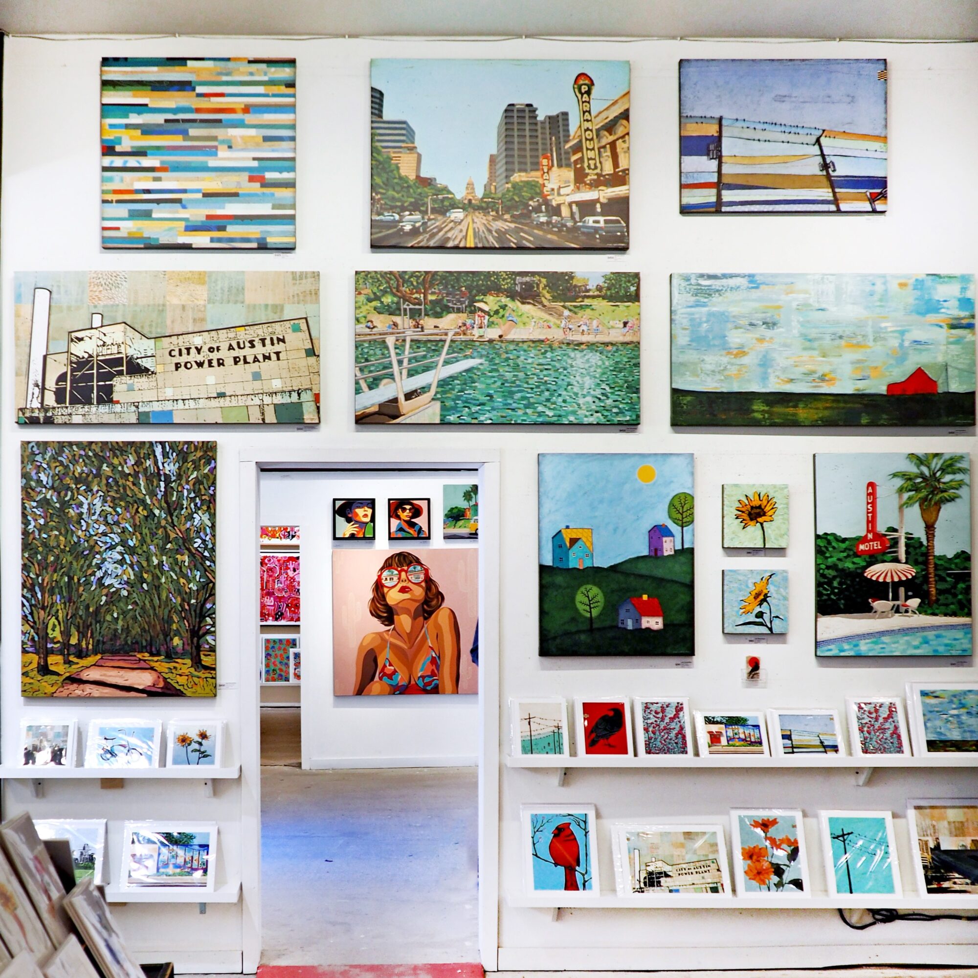 Gallery wall displaying framed paintings, photographs, and colorful artwork above a doorway and display shelves.