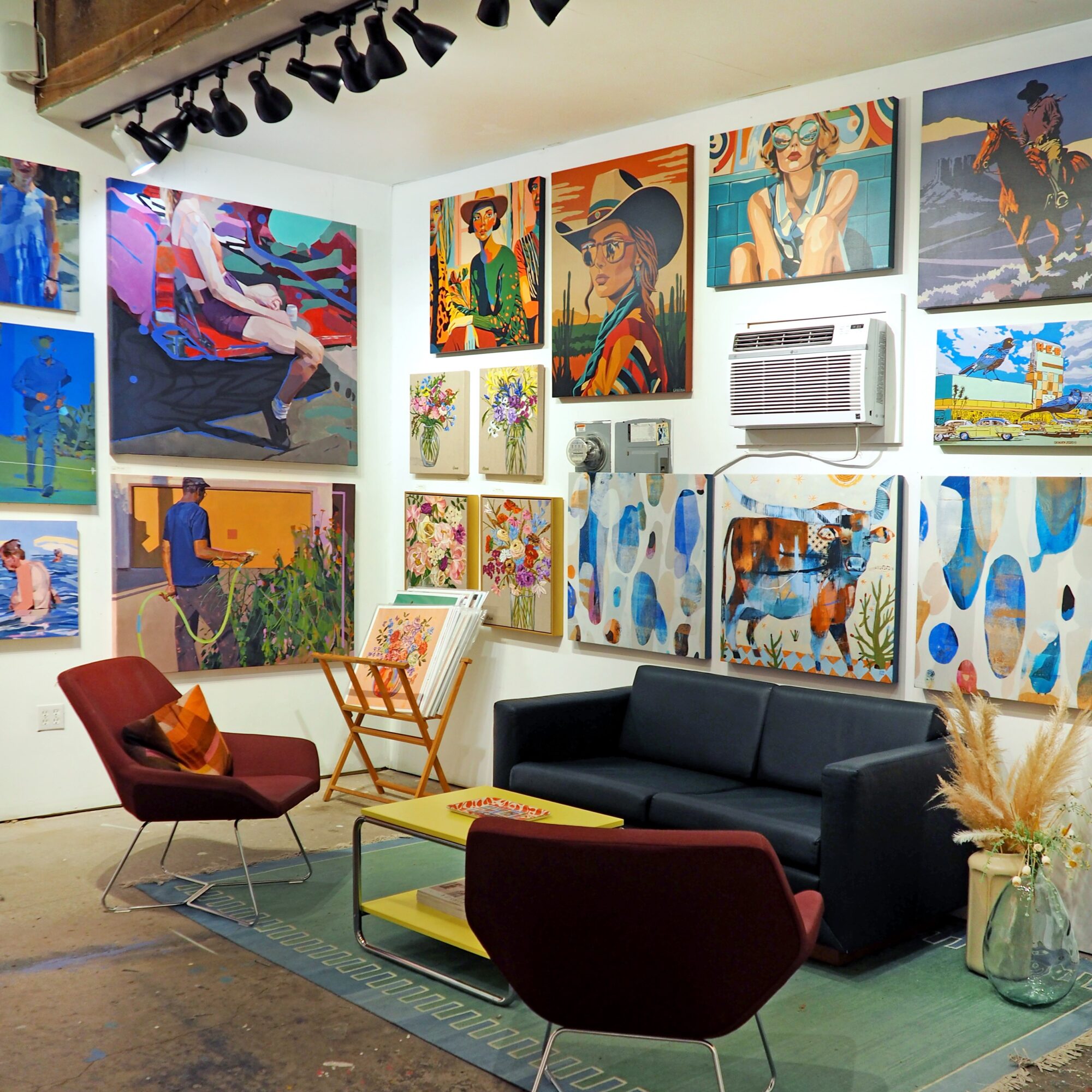 Gallery room with colorful paintings on the walls, a black sofa, two chairs, and a coffee table.
