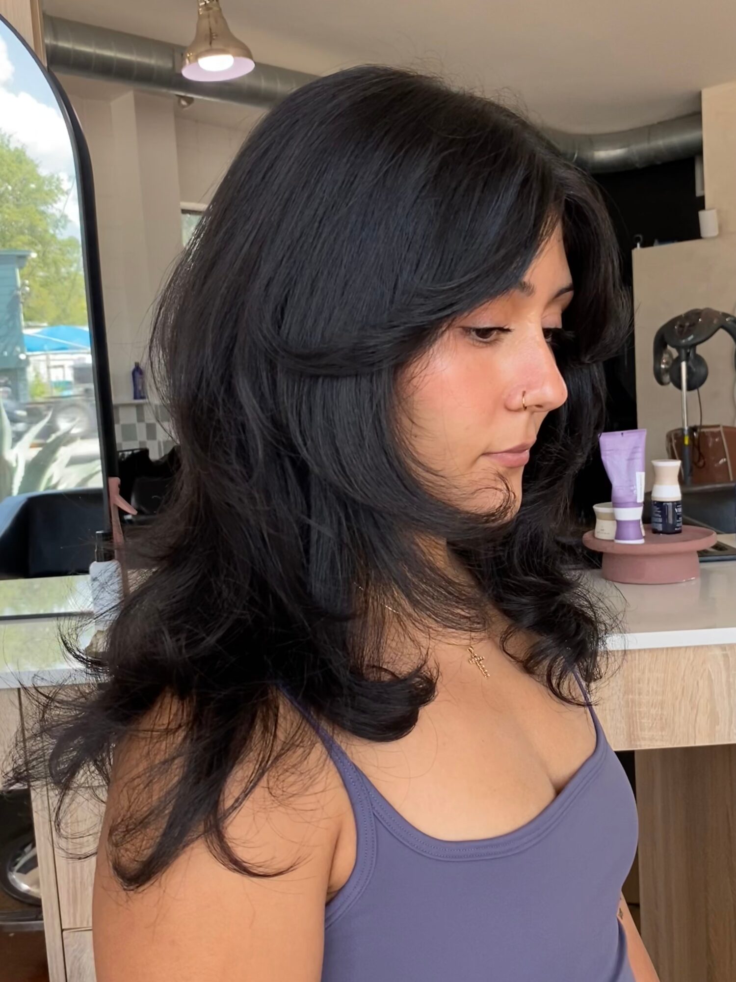 Person with long, layered dark hair wearing a purple top in a salon.