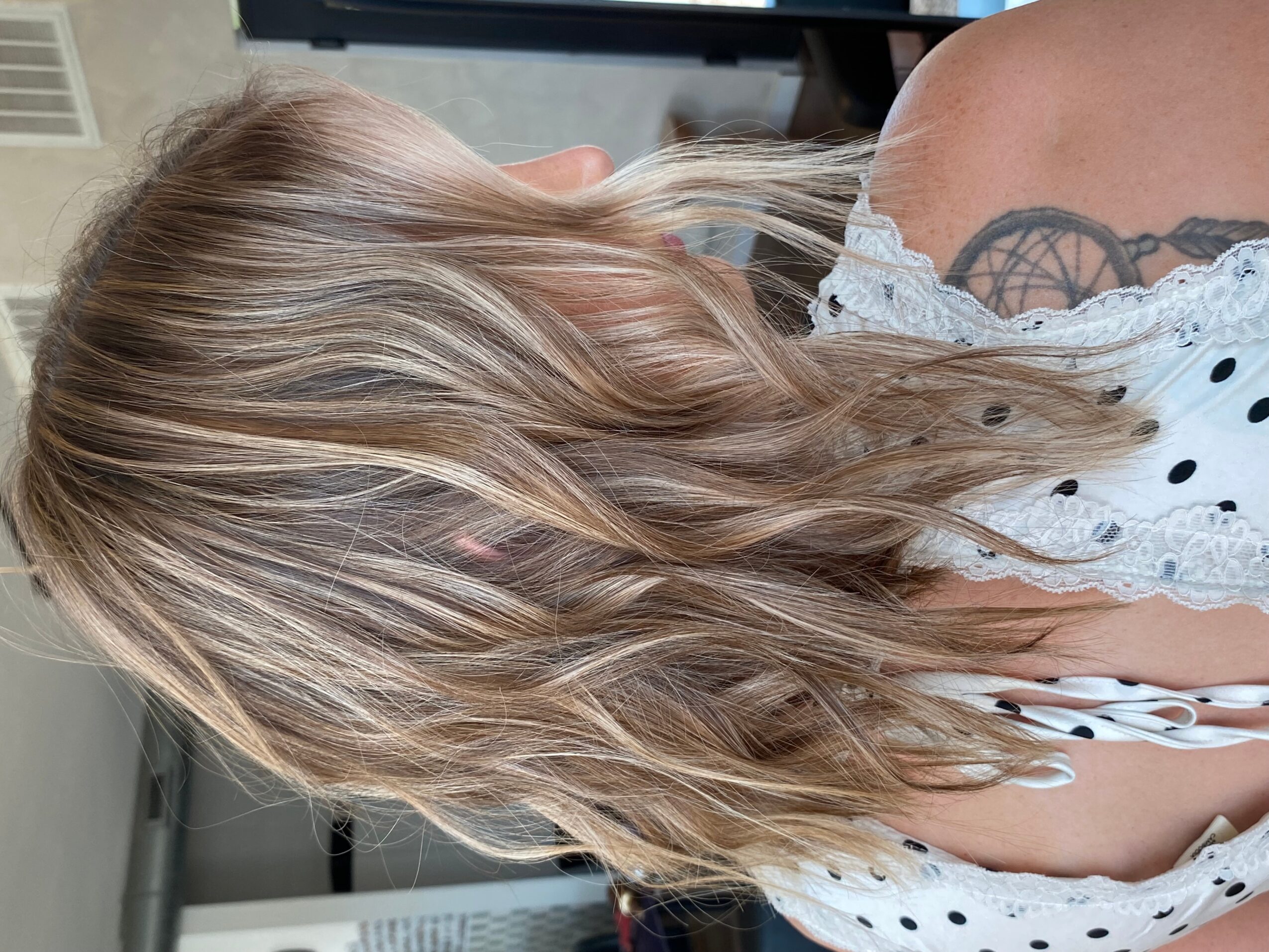 Person with long wavy hair wearing a white polka-dot top, with a tattoo visible on the upper arm.