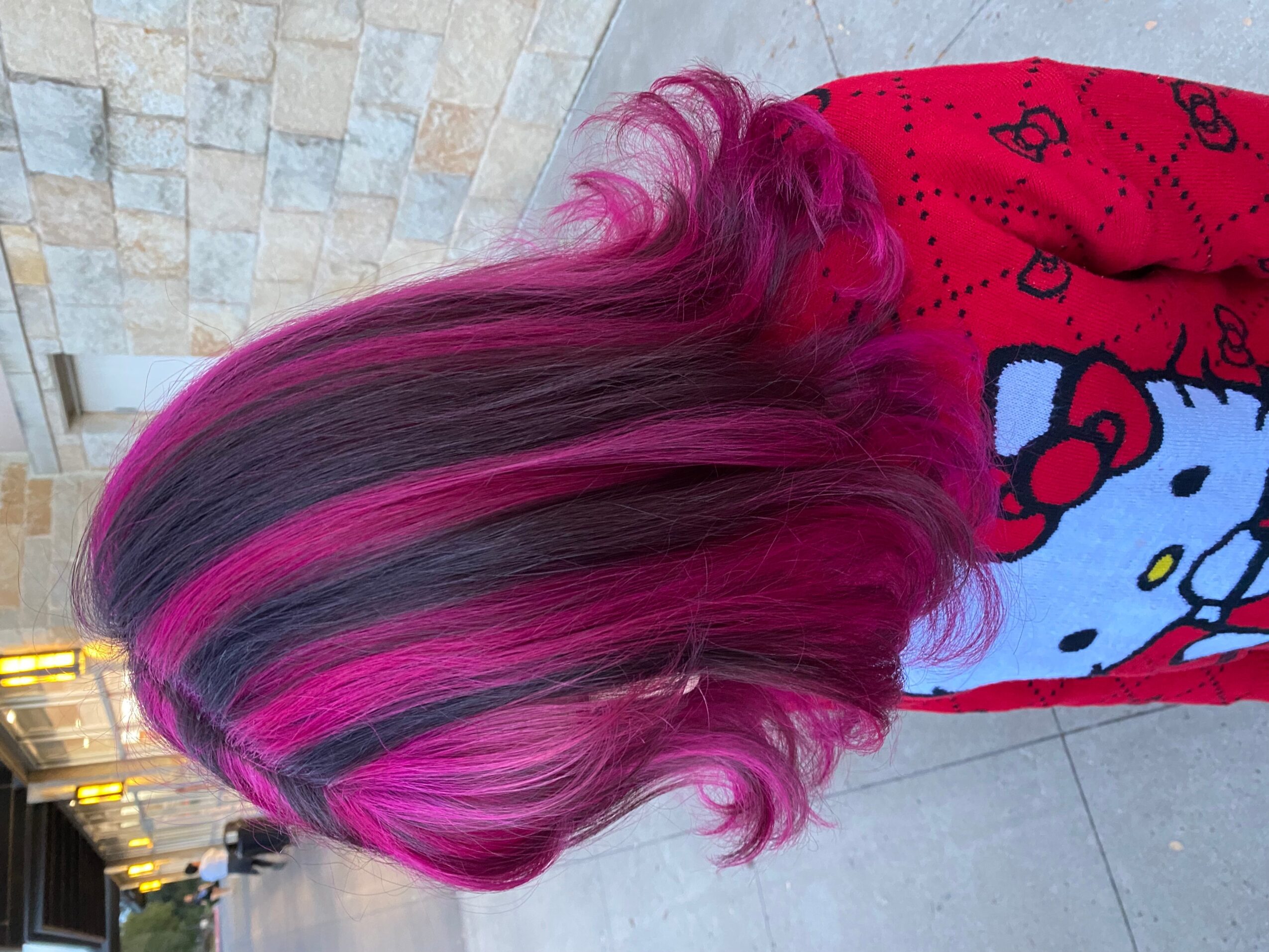 Person with long dark hair streaked with bright pink, wearing a red Hello Kitty shirt indoors.