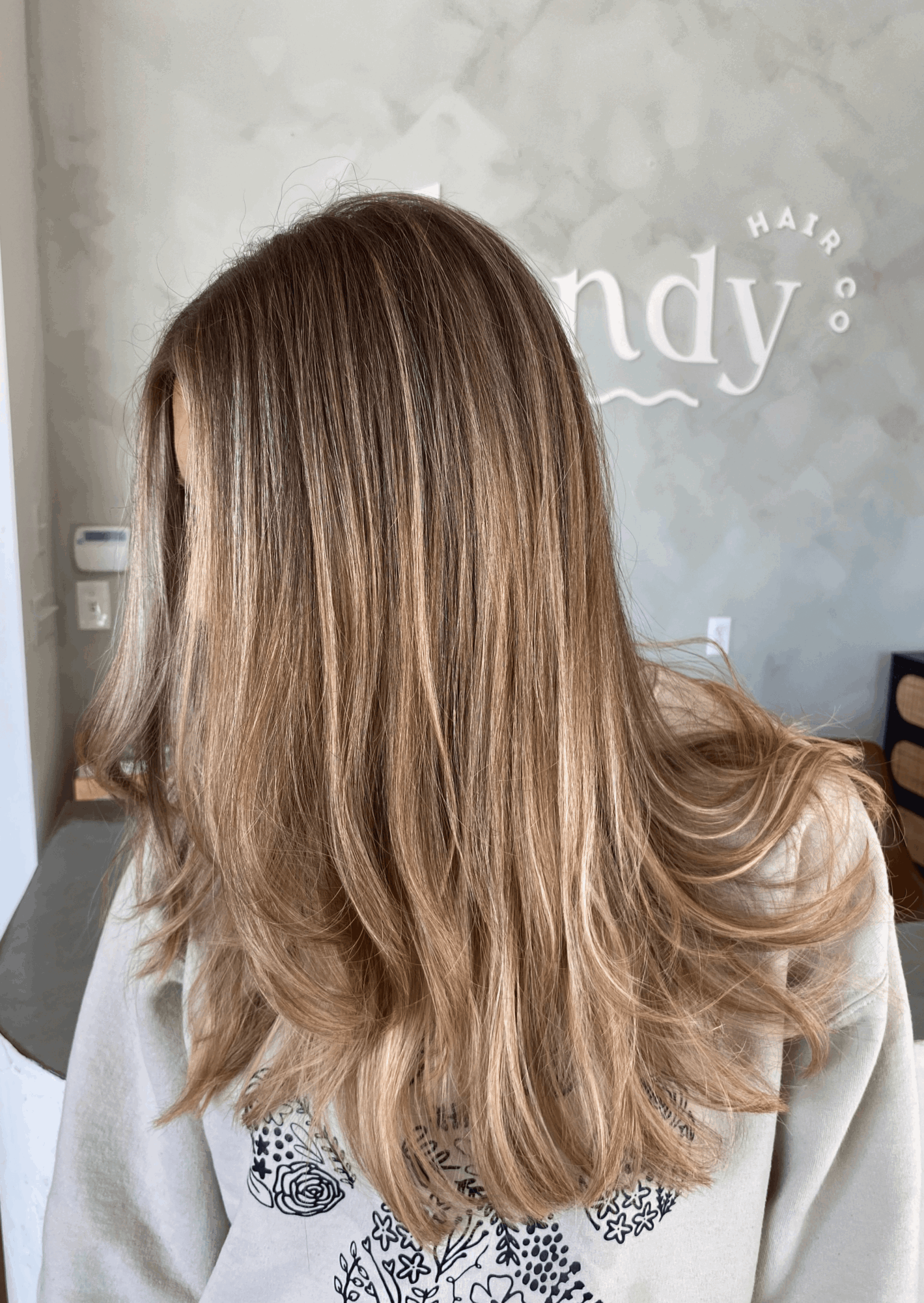 Person with long, layered light brown hair standing in front of a pale wall with partial lettering.