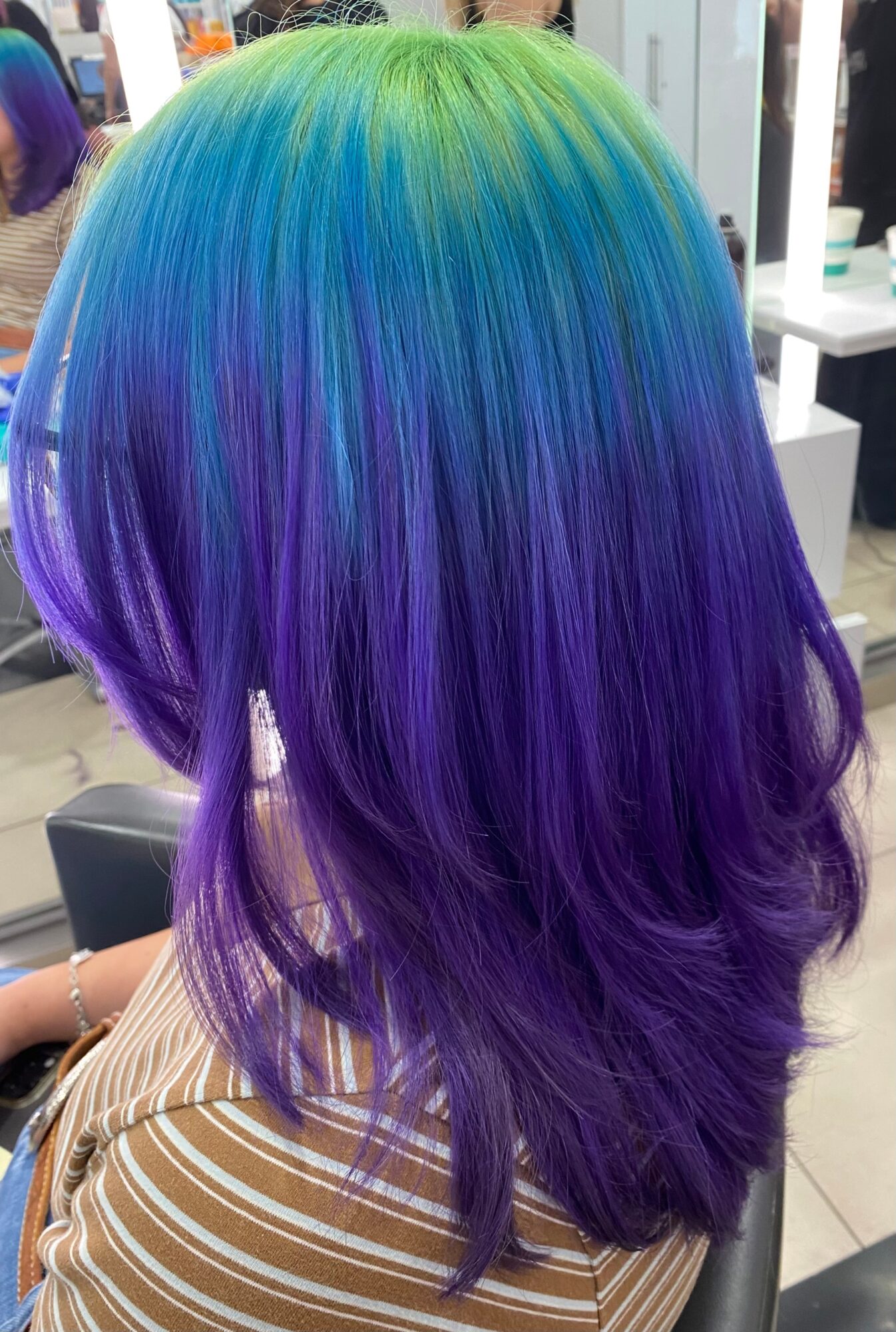 Person with long, wavy hair dyed green, blue, and purple, viewed from behind.