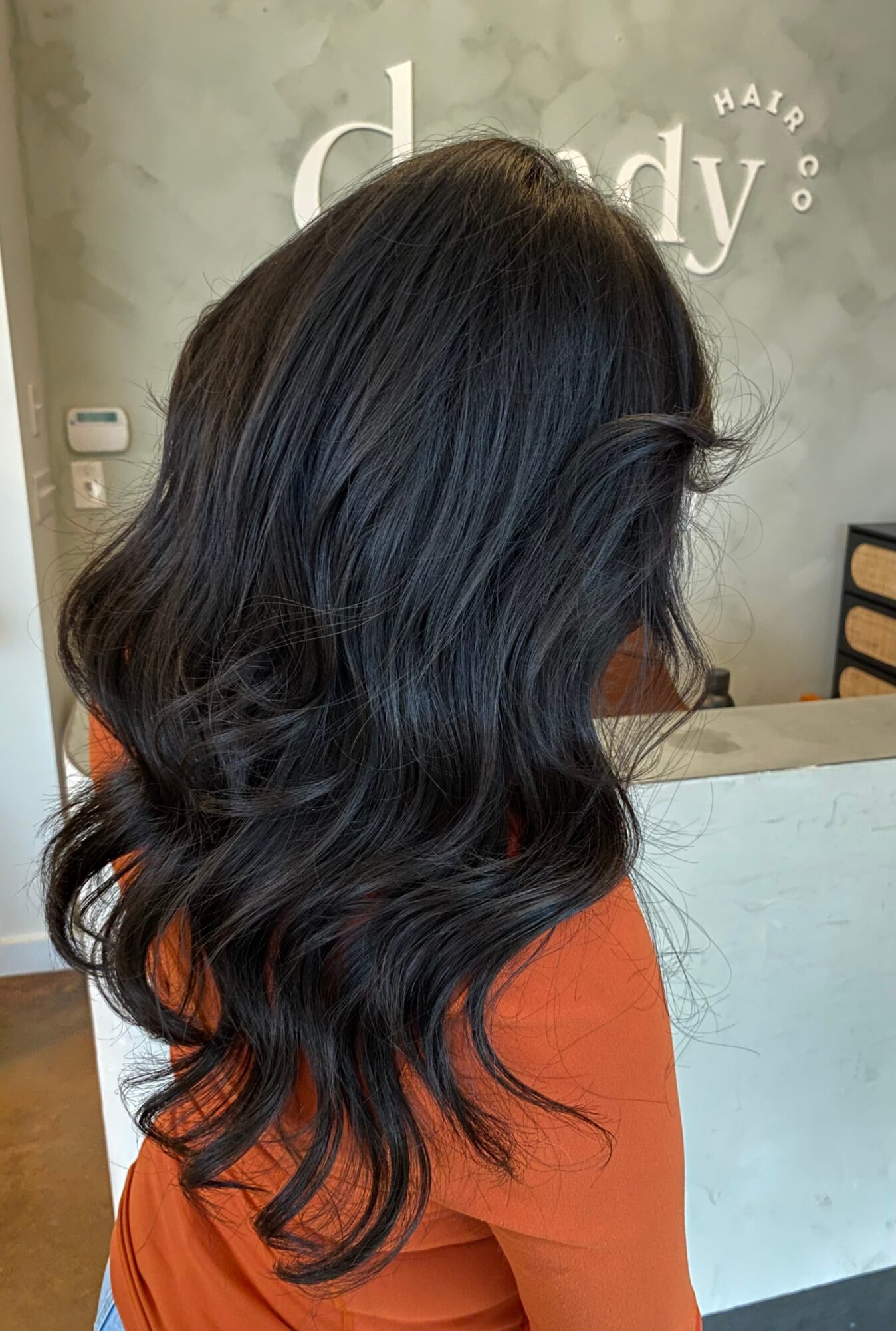 Person with long, wavy dark hair wearing an orange top in front of a salon counter.