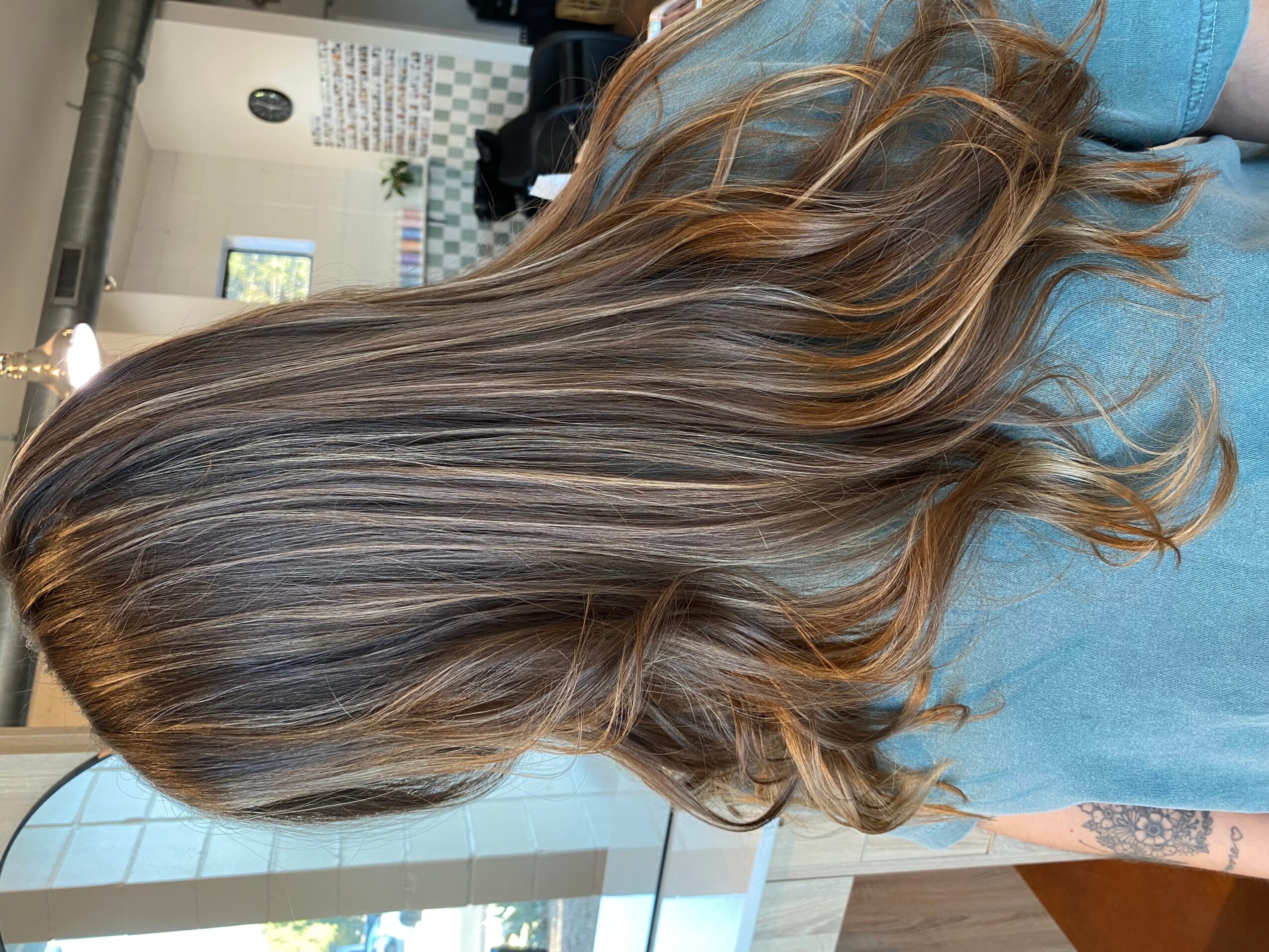 Long wavy brown hair with lighter highlights draped over a blue shirt in a salon.