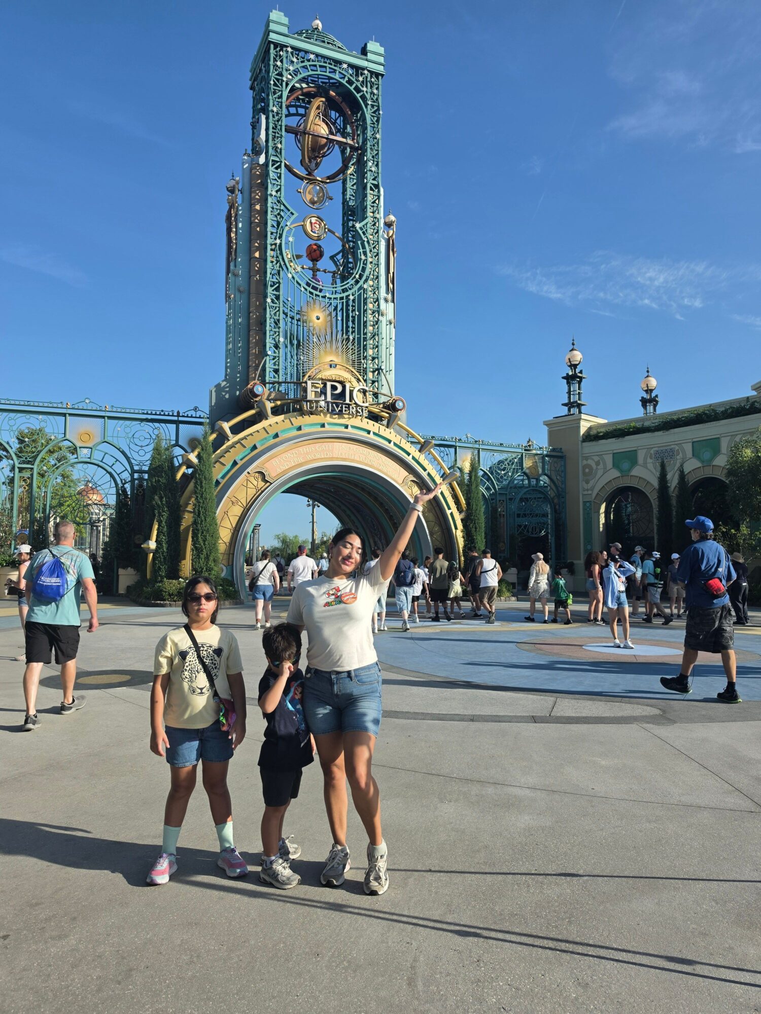 People stand beside an ornate green clock tower and arch in a sunny plaza.