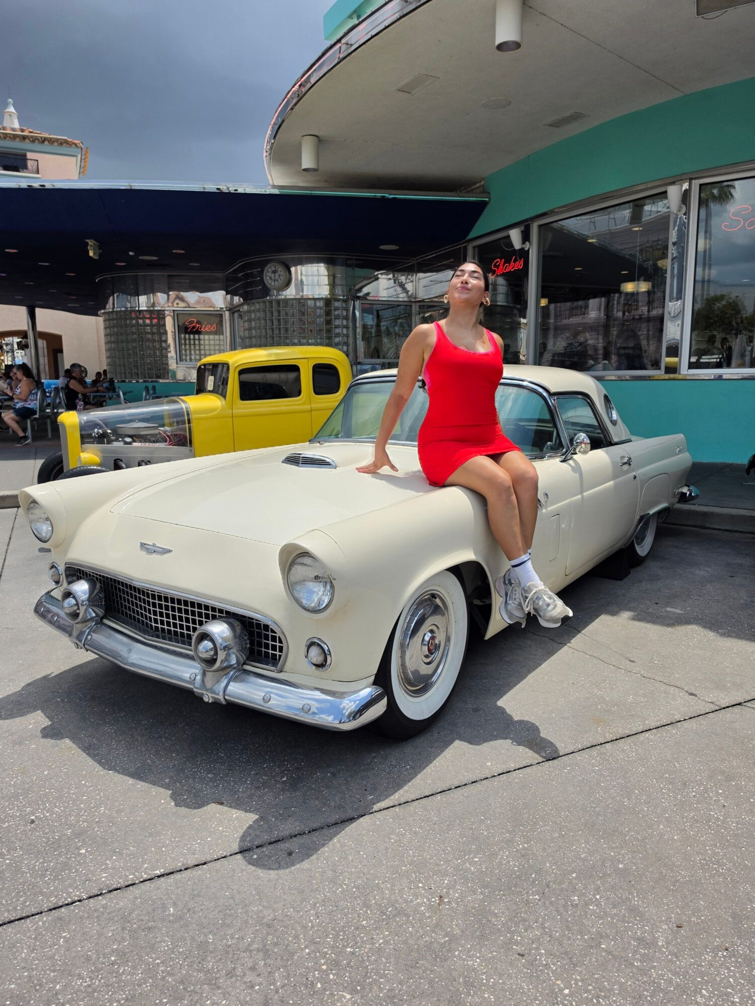 Person in a red top standing beside a white vintage car outside a building, with a yellow vehicle in the background.
