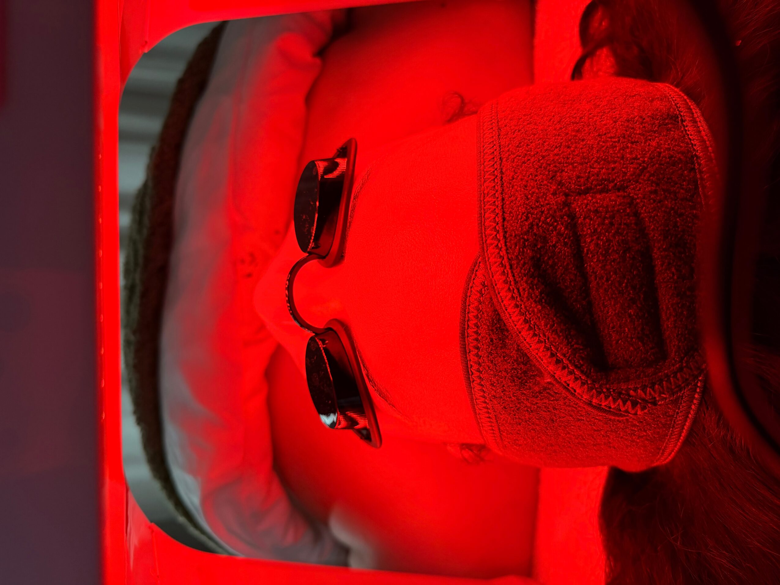 Close-up of a person wearing dark sunglasses and a textured face covering under red lighting.