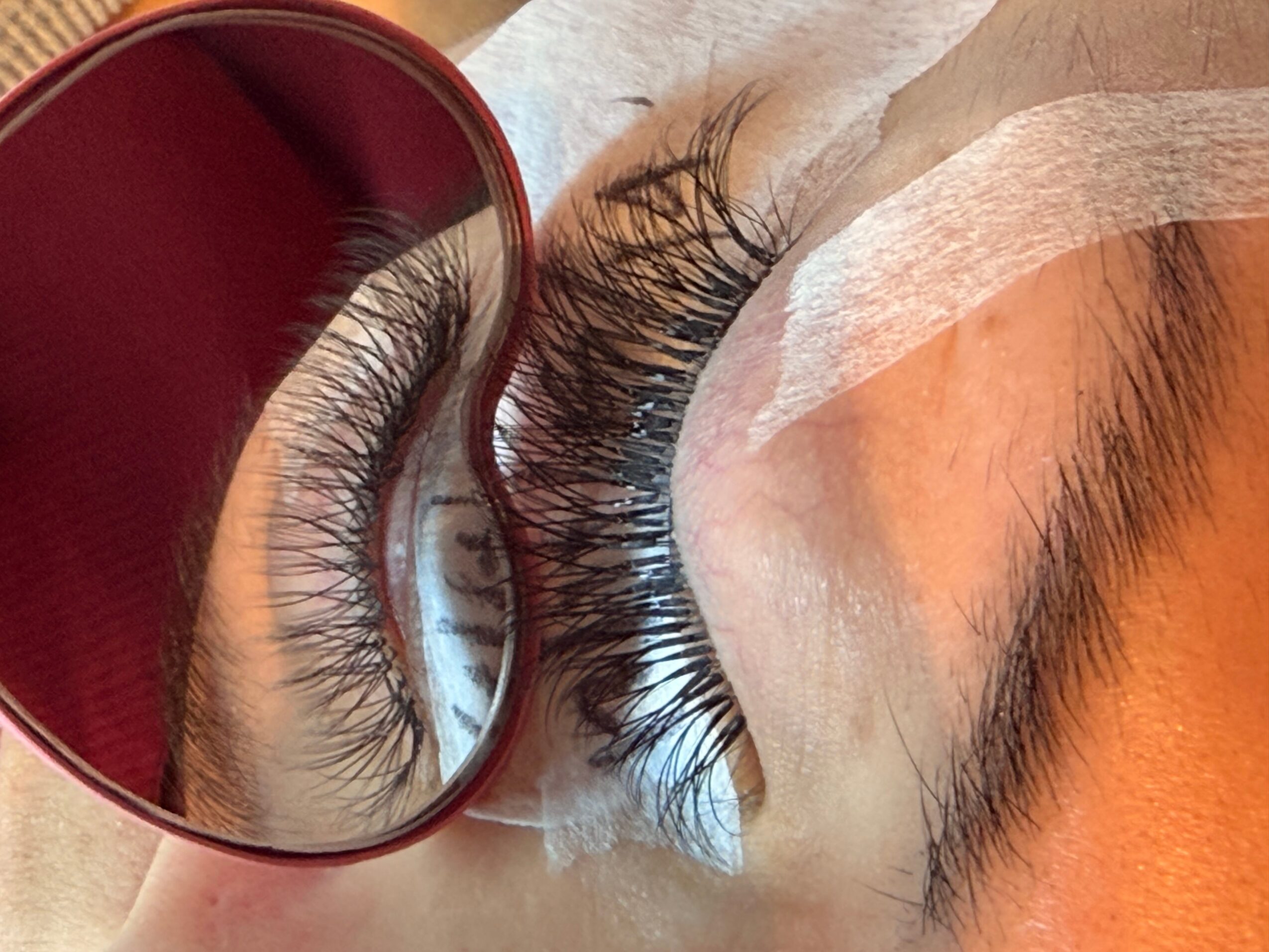 Close-up of a person’s closed eye with long eyelashes and a red-rimmed mirror nearby.