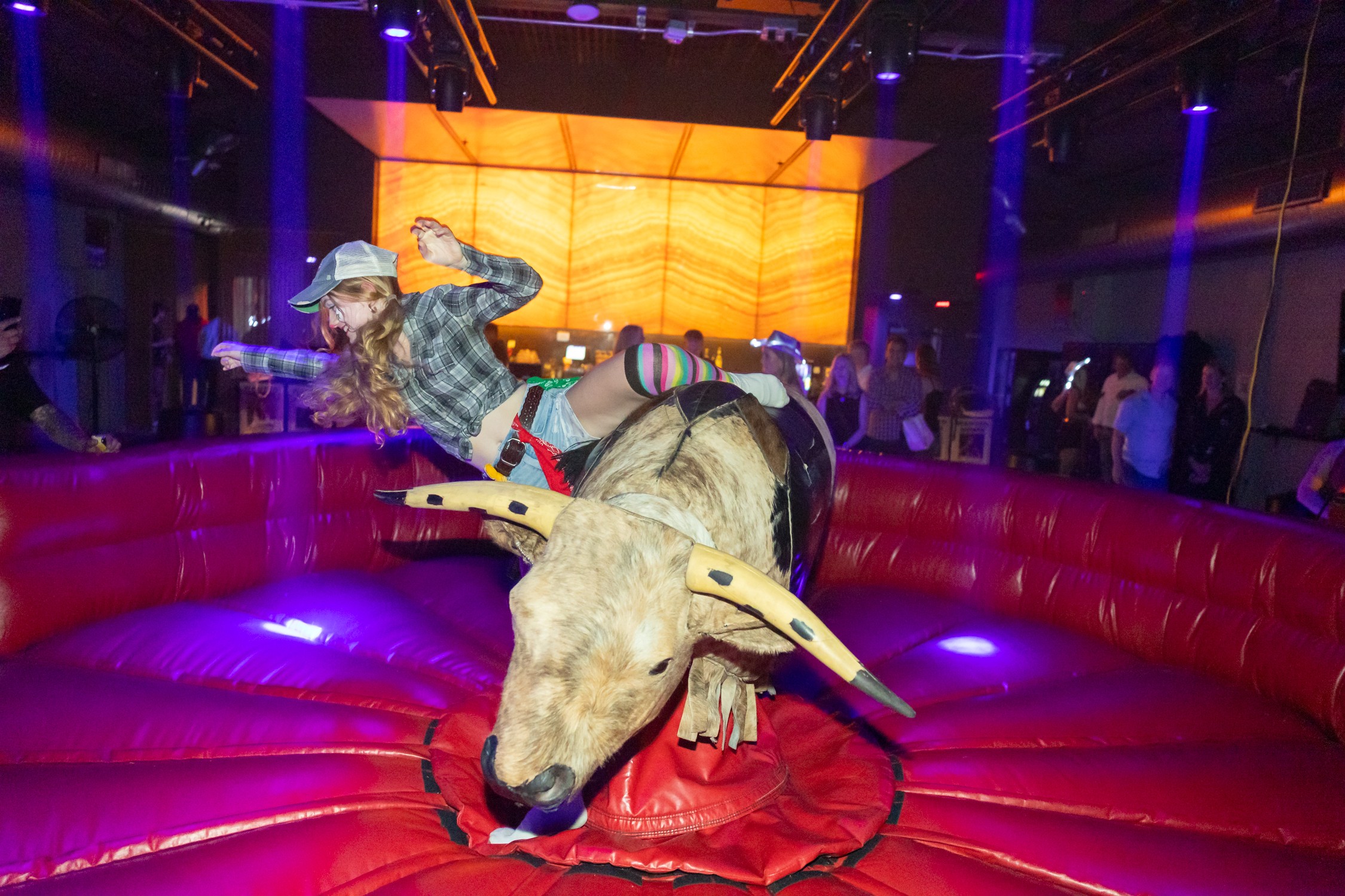 A person in a cowboy hat rides a mechanical bull on a padded arena floor as spectators watch.