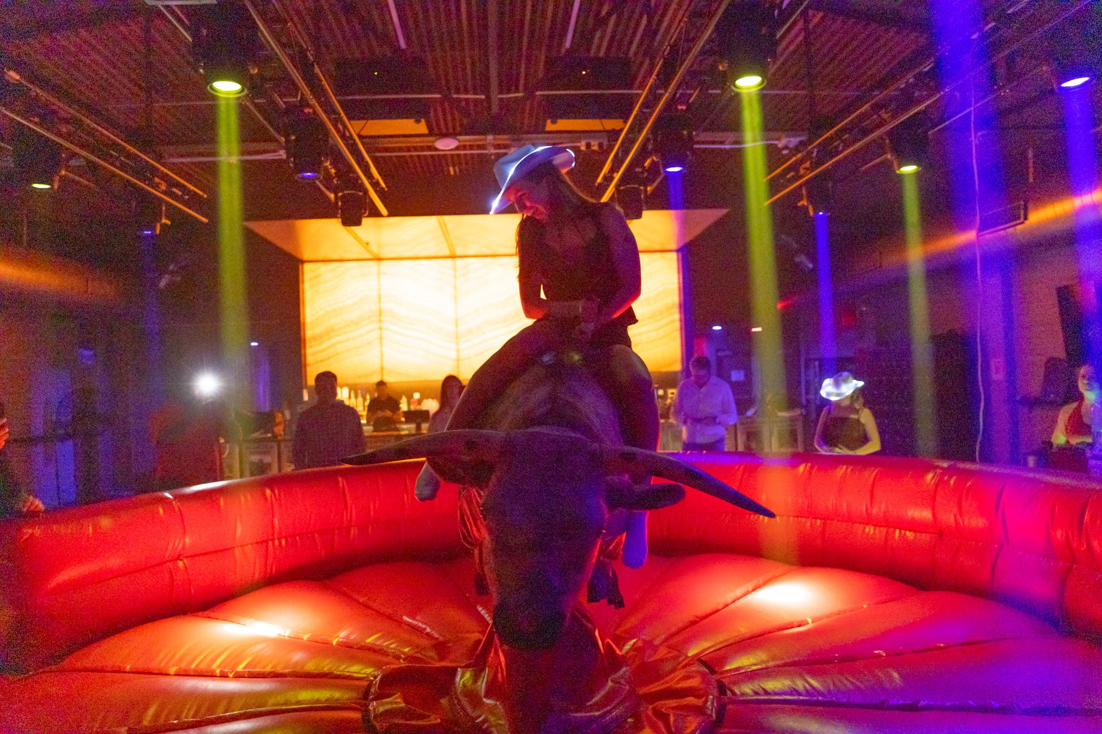 A person rides a mechanical bull in a red inflatable arena under colorful stage lights.