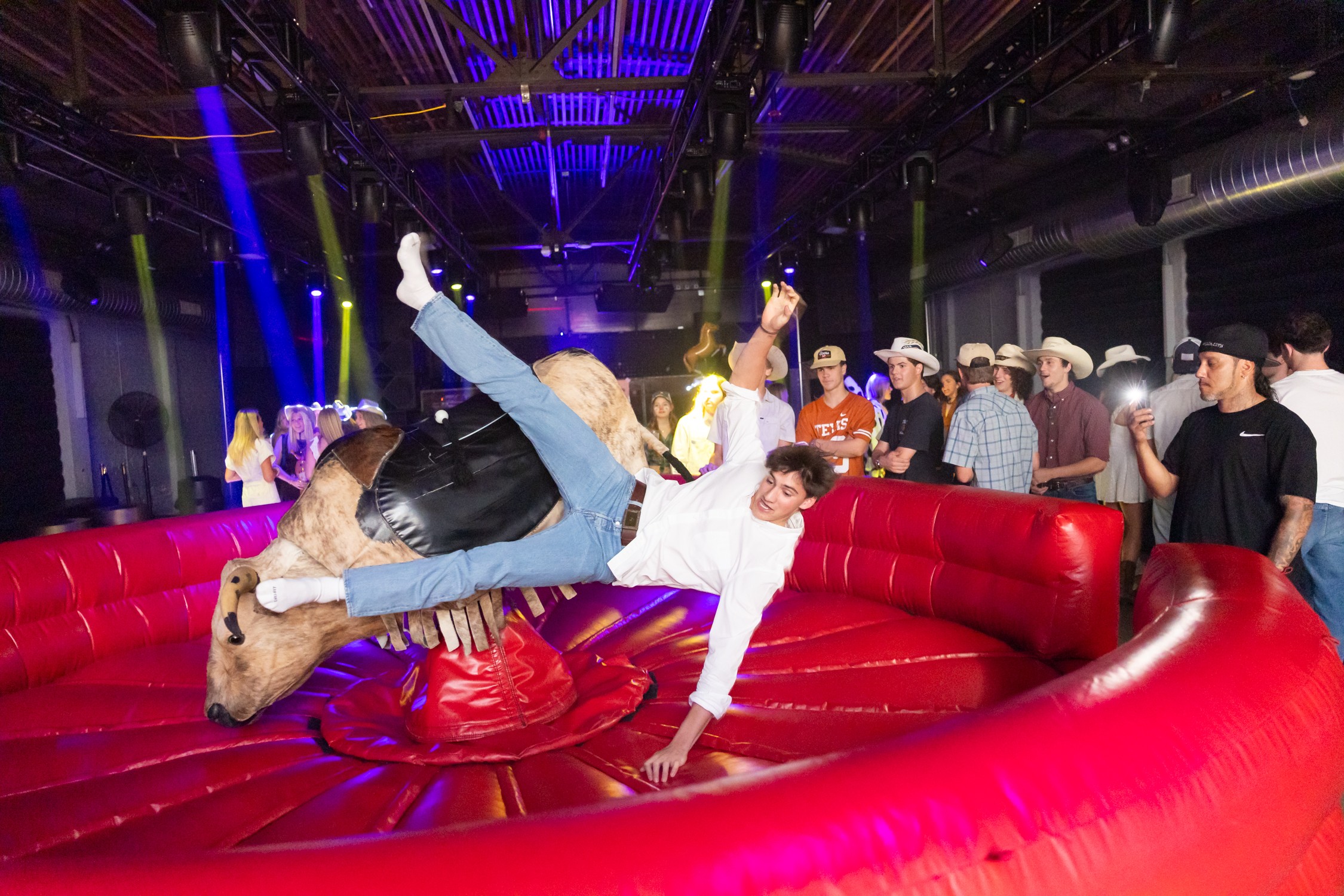A person in a white shirt and jeans falls across a red mechanical bull ring as spectators watch.