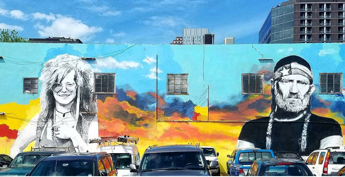 Colorful mural of two large monochrome portraits above parked cars, with city buildings and blue sky behind.