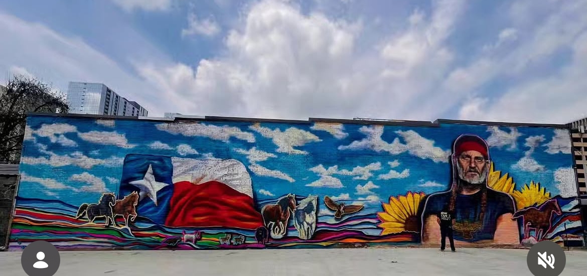 Long colorful mural depicting a flag, desert landscape, sun, and a person against a blue sky with clouds.
