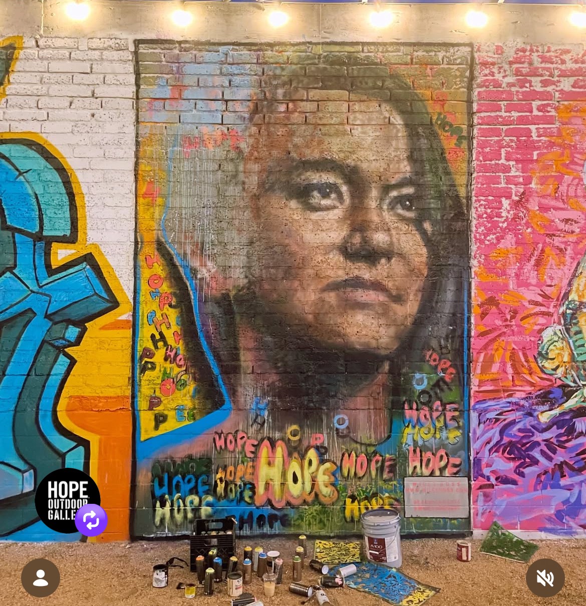 Large painted portrait of a person on a brick wall, surrounded by colorful graffiti and paint supplies.