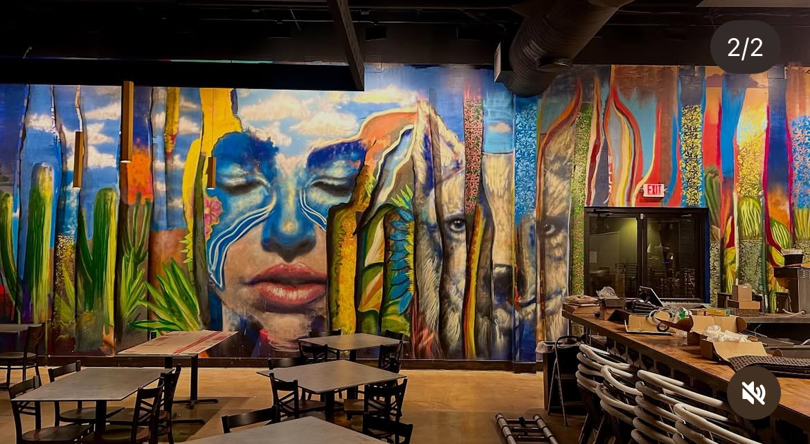 Restaurant dining area with tables and chairs before a colorful mural featuring a large face and abstract patterns.