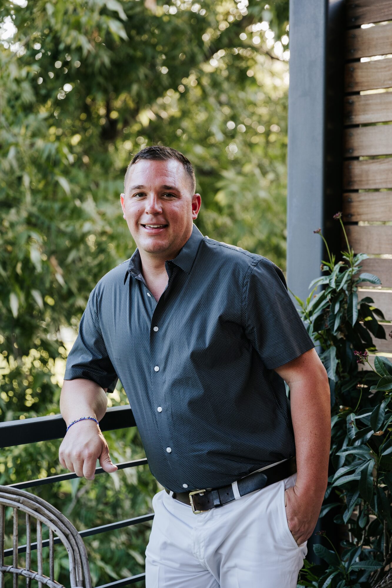 A person in a dark shirt and white trousers stands beside a wooden wall amid leafy plants.