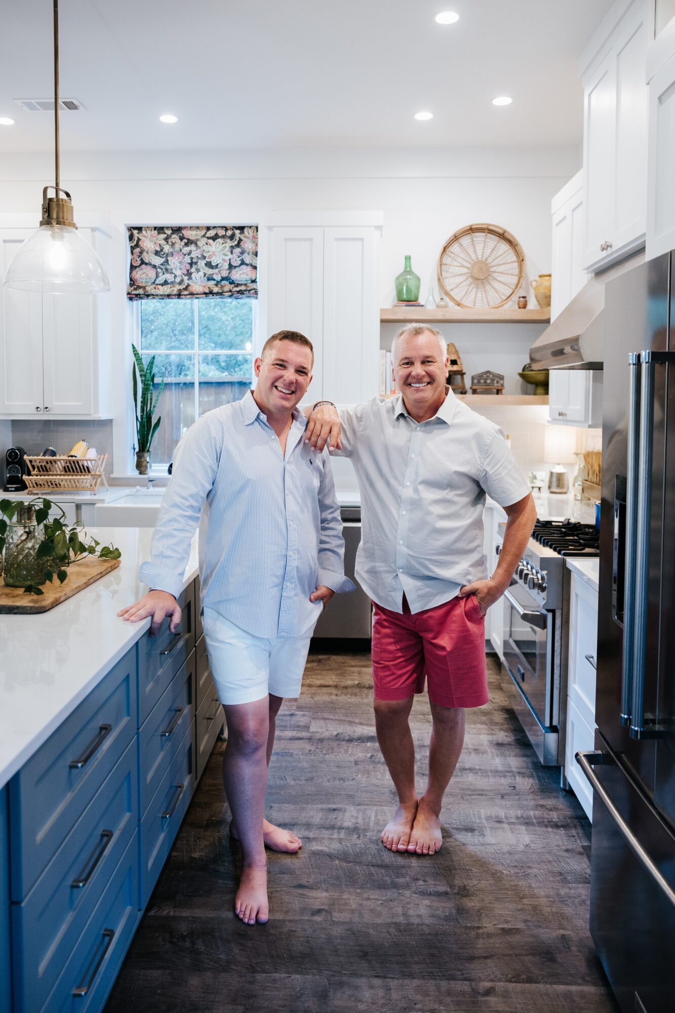 Two people stand arm in arm in a bright kitchen with blue cabinets and a large window.