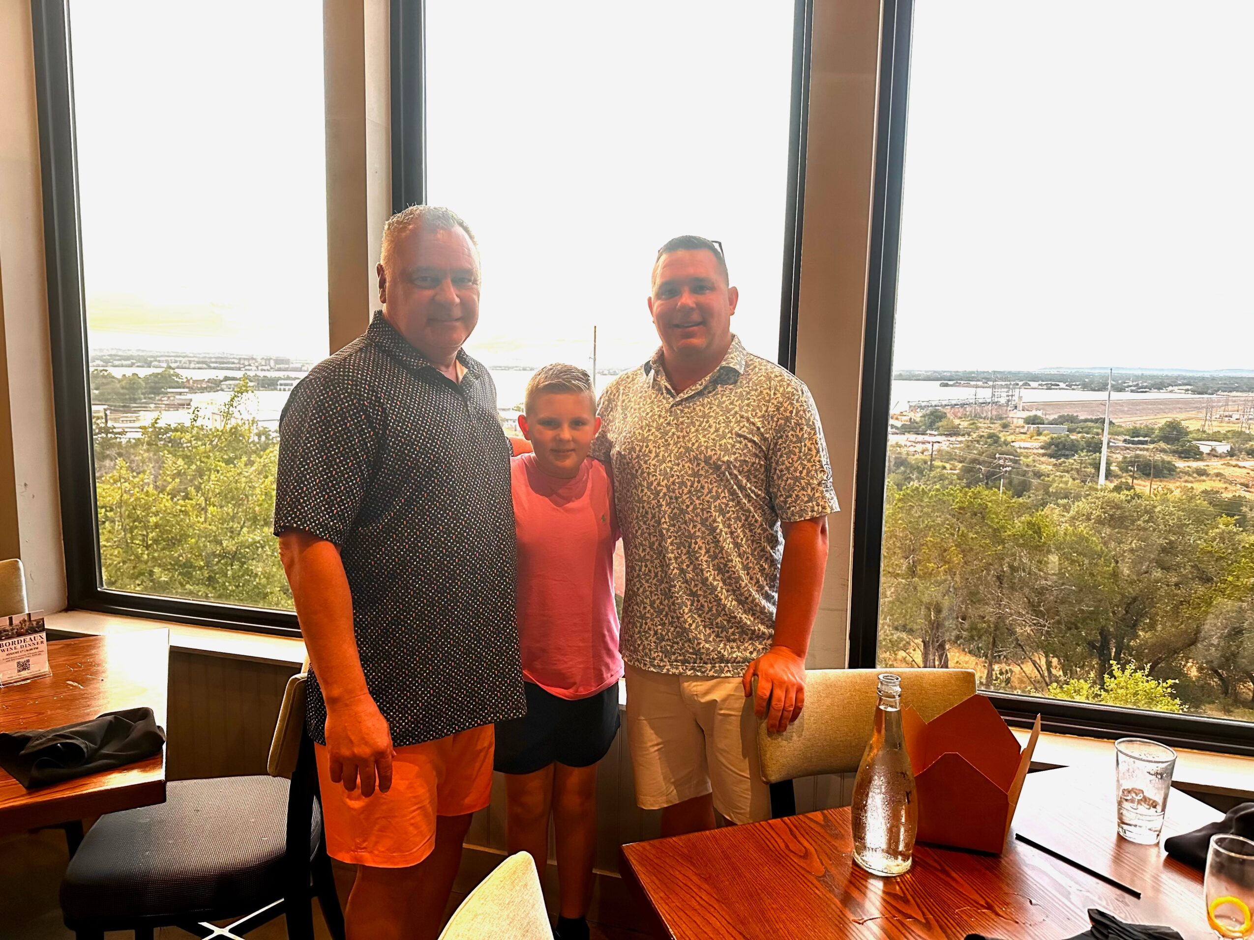 Two adults and a child stand beside a dining table in front of large windows overlooking a landscape.
