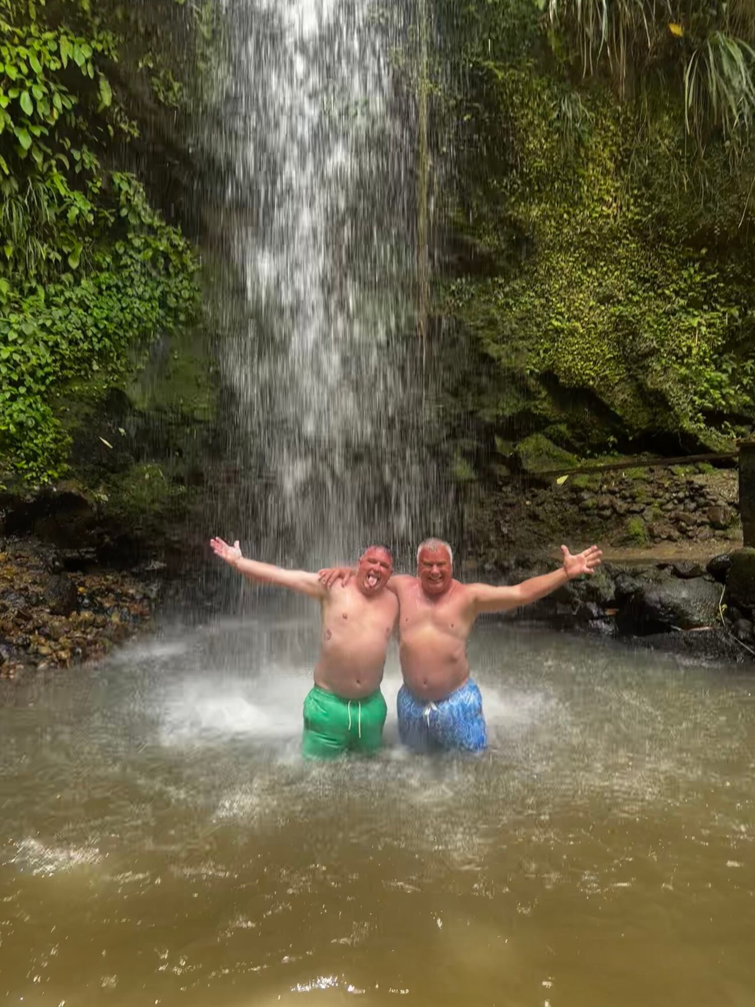Two people stand in a pool beneath a waterfall with their arms outstretched.