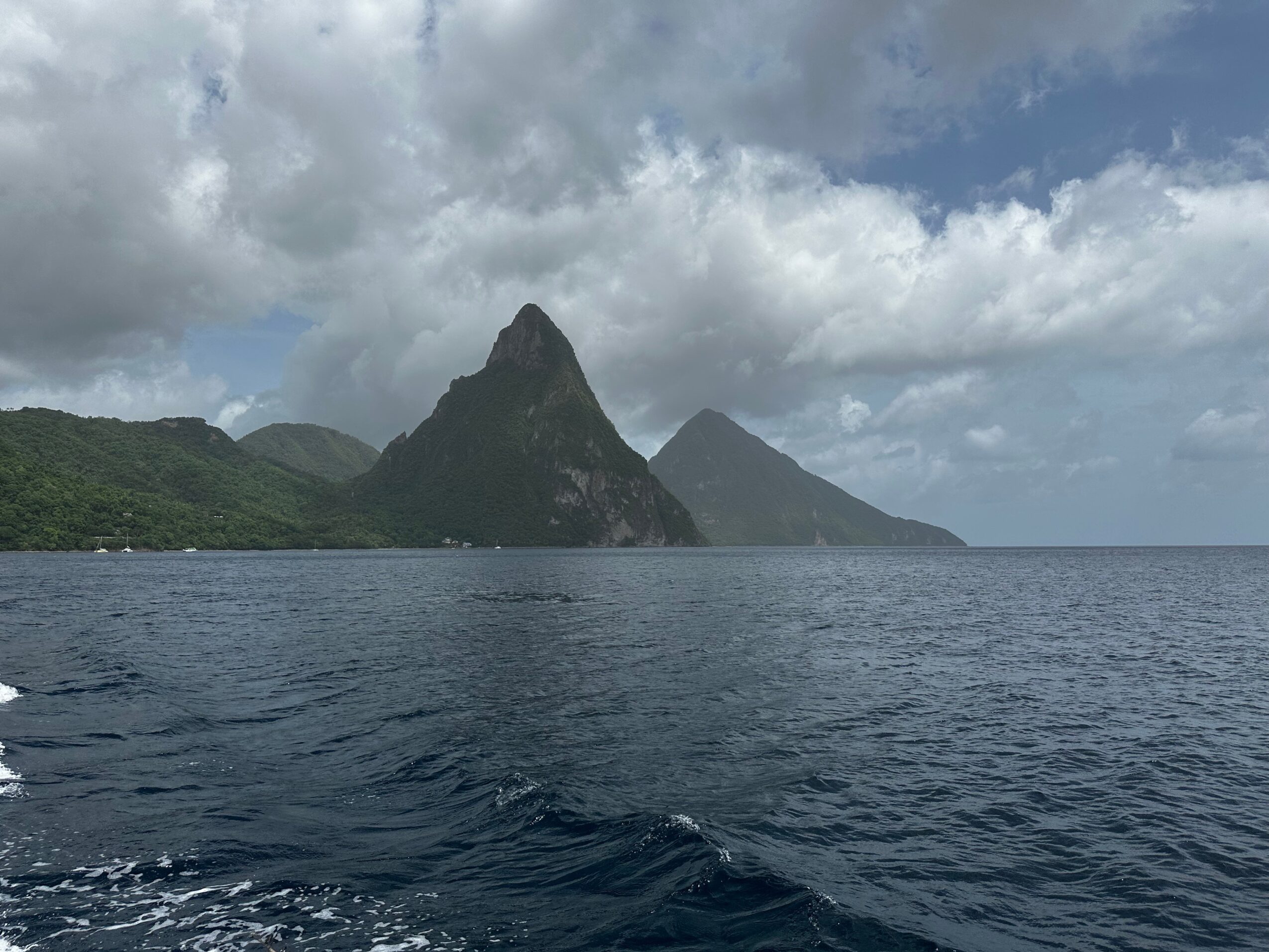 Mountain peaks rise above a forested coastline across dark water beneath cloudy skies.