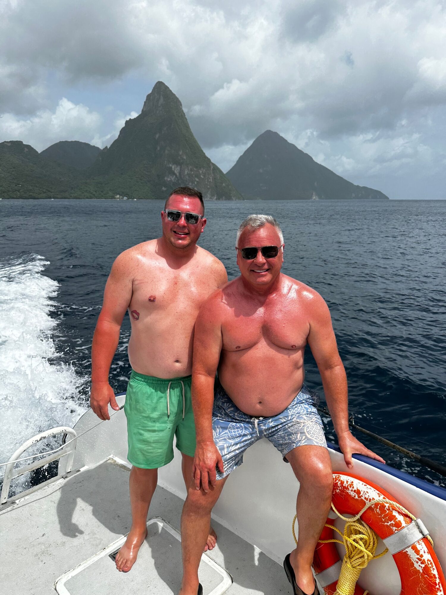 Two people in swimwear aboard a boat, with mountains, water, and cloudy skies behind them.