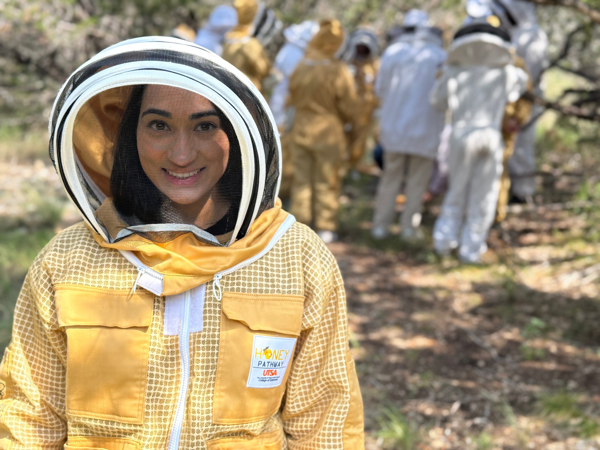 Person wearing a yellow beekeeping suit and protective hood, with several suited people among trees in the background.