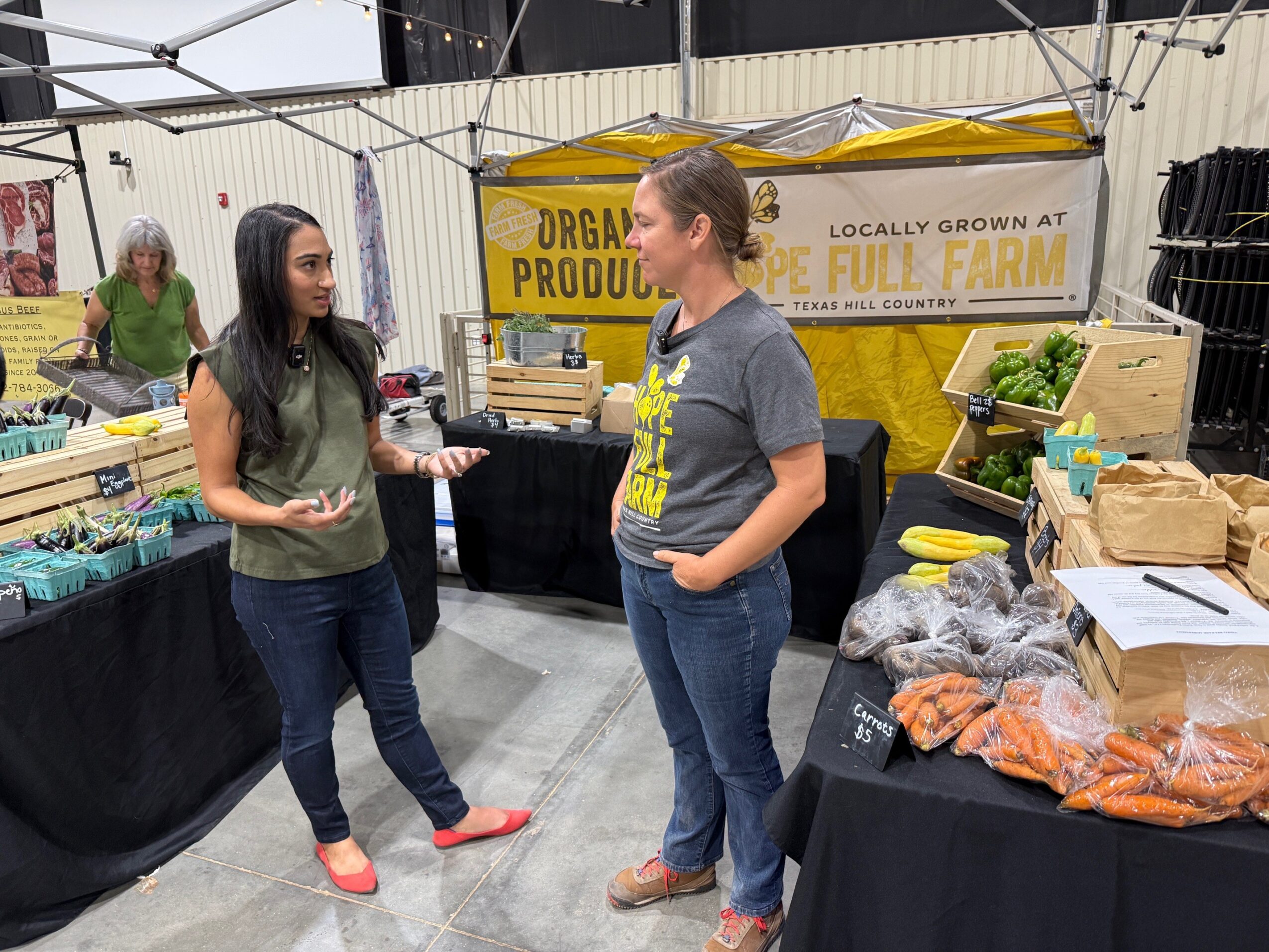 Two people talk beside tables displaying packaged produce at an indoor market booth.