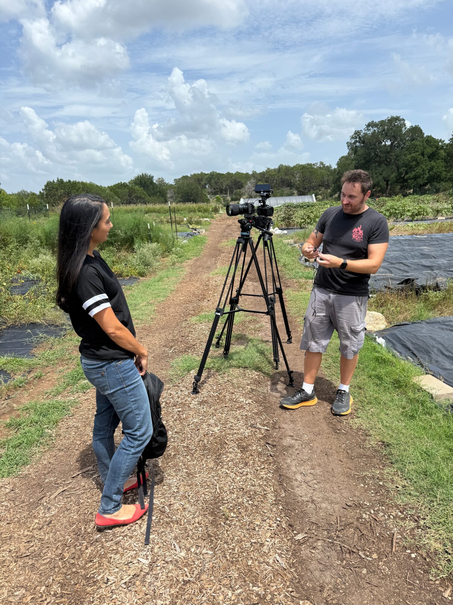 Two people stand beside a tripod camera on a dirt path near a grassy field.