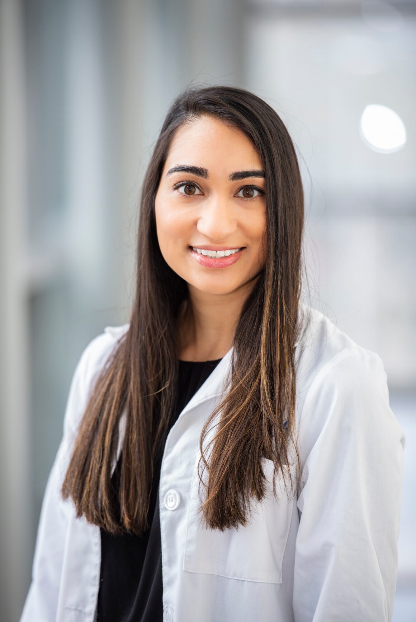 A person with long dark hair wearing a white lab coat over a black top, standing indoors.