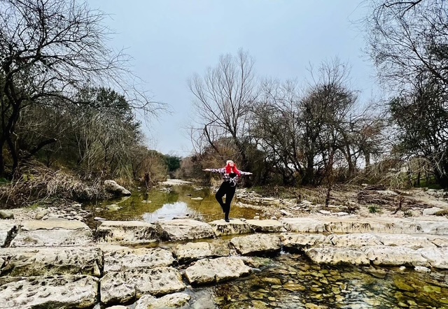 Person standing on pale rocky ground in a shallow stream, surrounded by leafless trees.